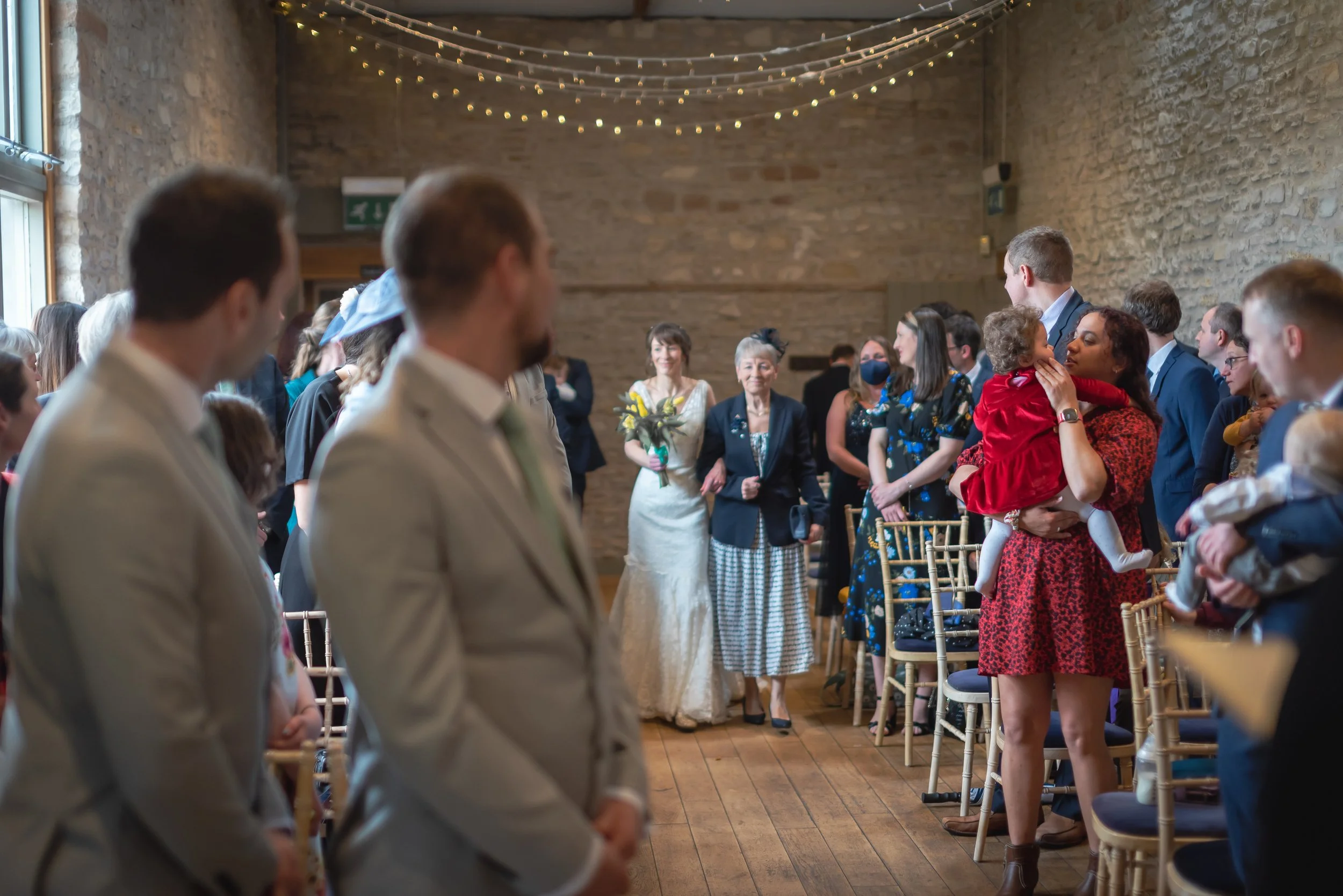 People gathered at a wedding ceremony in a rustic venue with string lights and exposed brick walls. The bride in a white dress holds a bouquet, and the groom is out of focus in the foreground. Guests are standing, some holding children, and appear emotional.