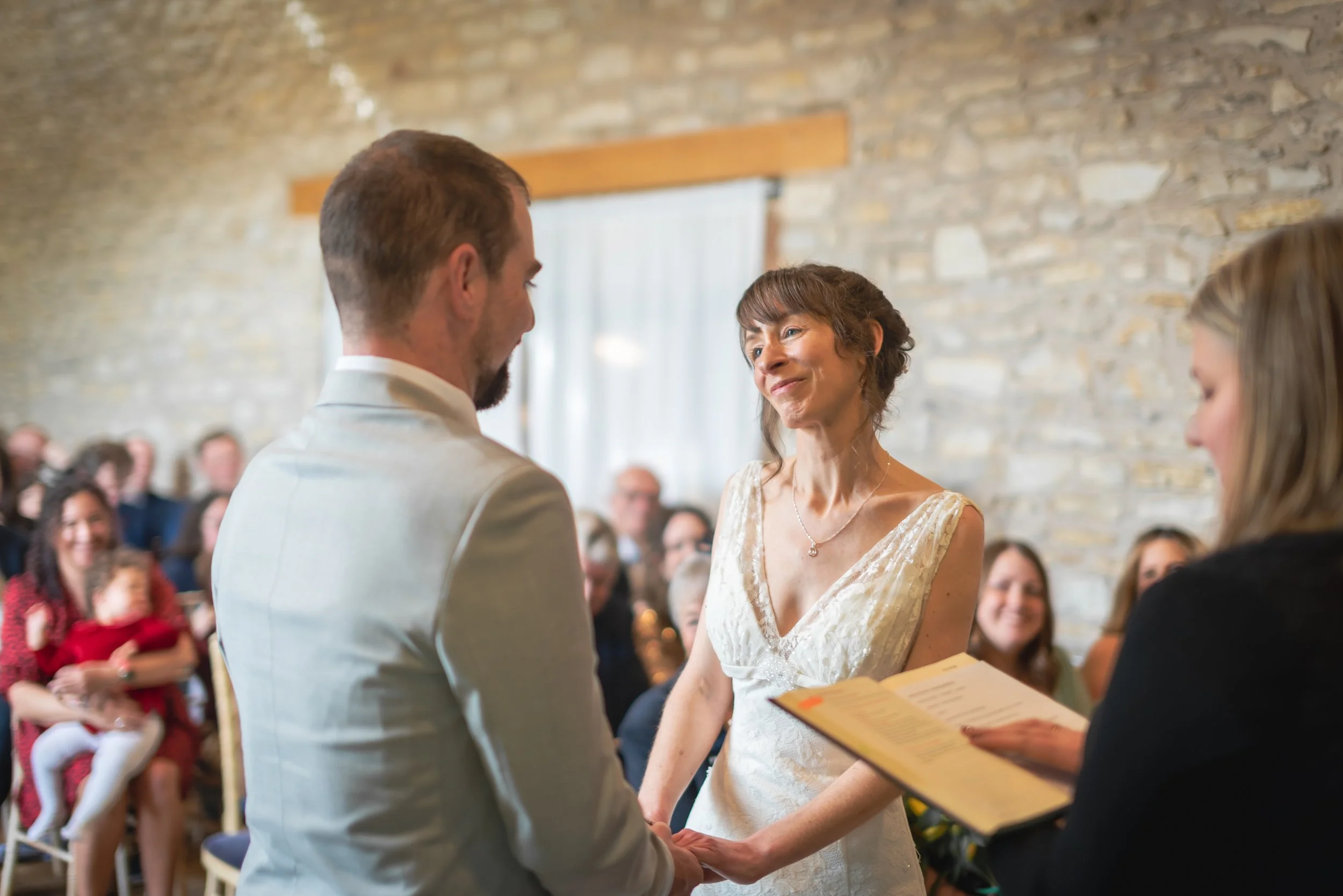 Folly Farm Wedding Photographer captures a bride and groom holding hands during a wedding ceremony, exchanging vows in front of an officiant, with guests seated in the background.