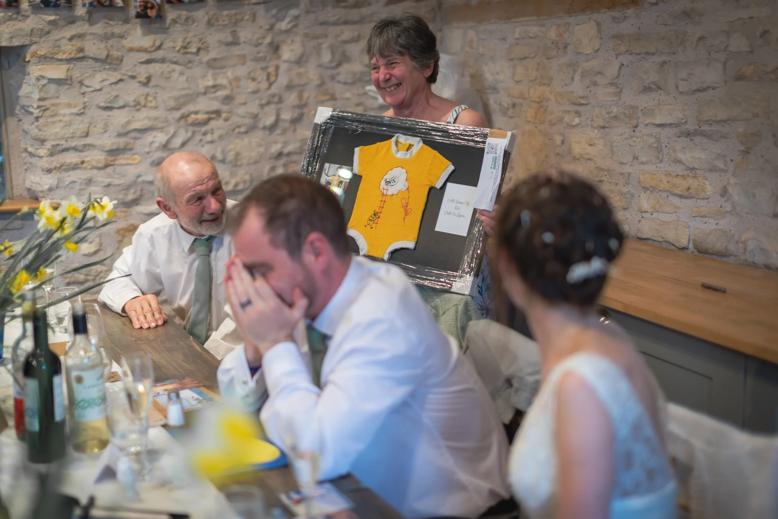 A bride smiling and holding a framed handmade yellow T-shirt with a drawing, at a celebration with three people sitting at a table, groom and parents with drinks and flowers at Folly Farm.