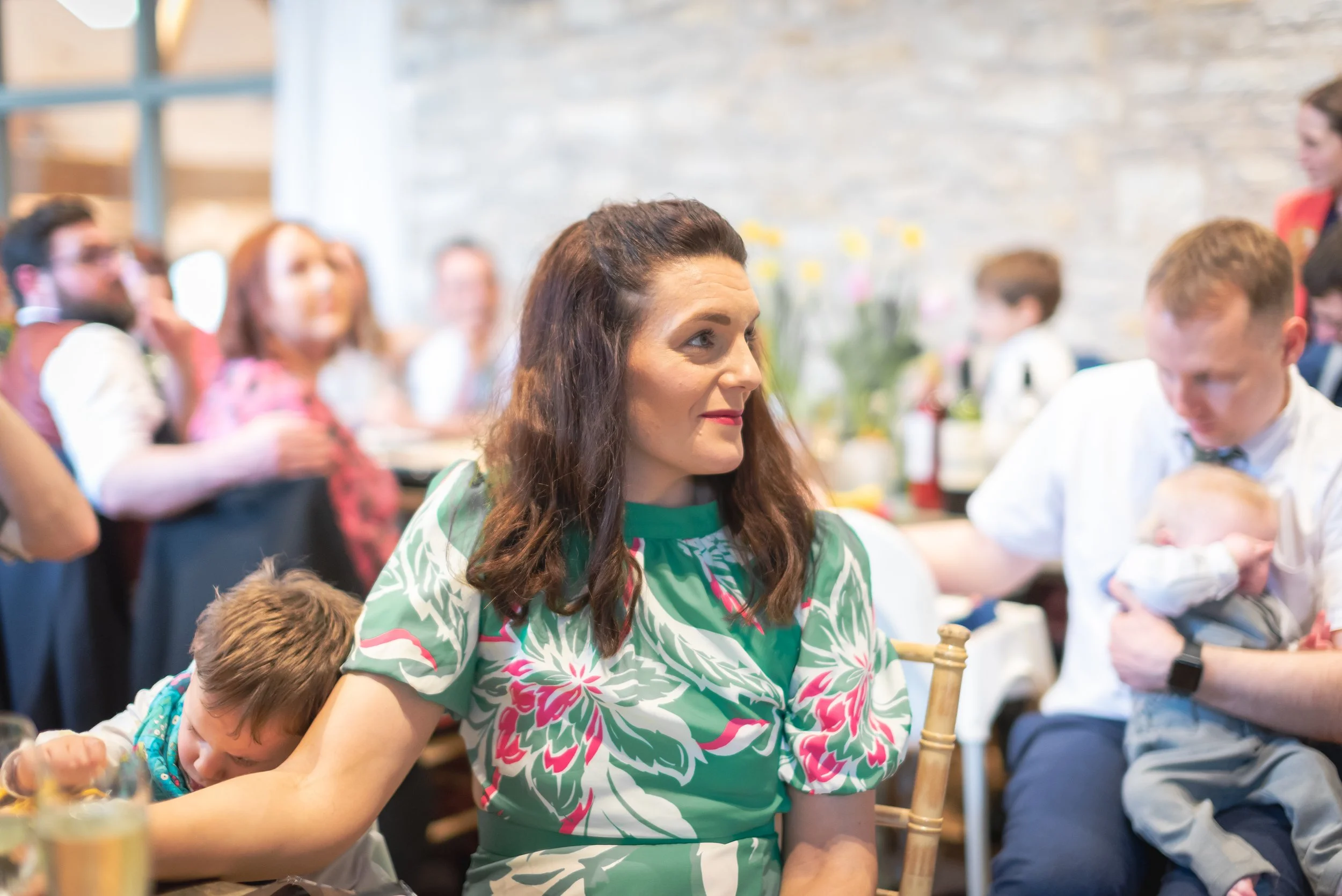 A woman in a green floral dress sitting at a table during a social gathering, with children and adults around her, some holding babies, in a bright room with stone walls.