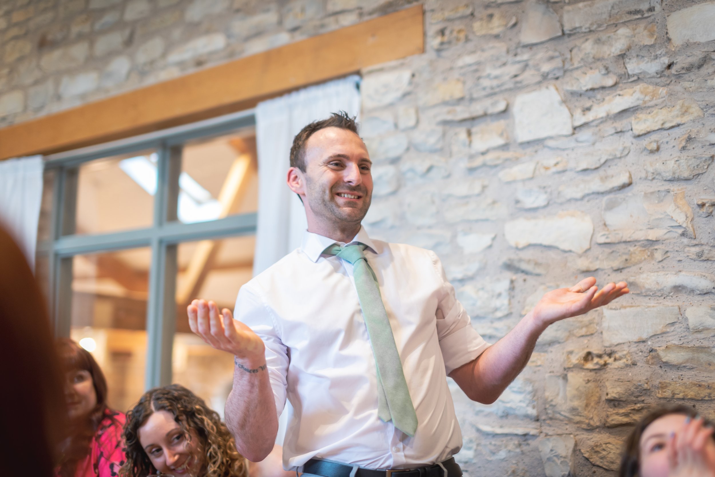 Best man in a white shirt and light green tie smiling with his arms raised in a room with a stone wall and large window captured by Bristol Wedding Photographer