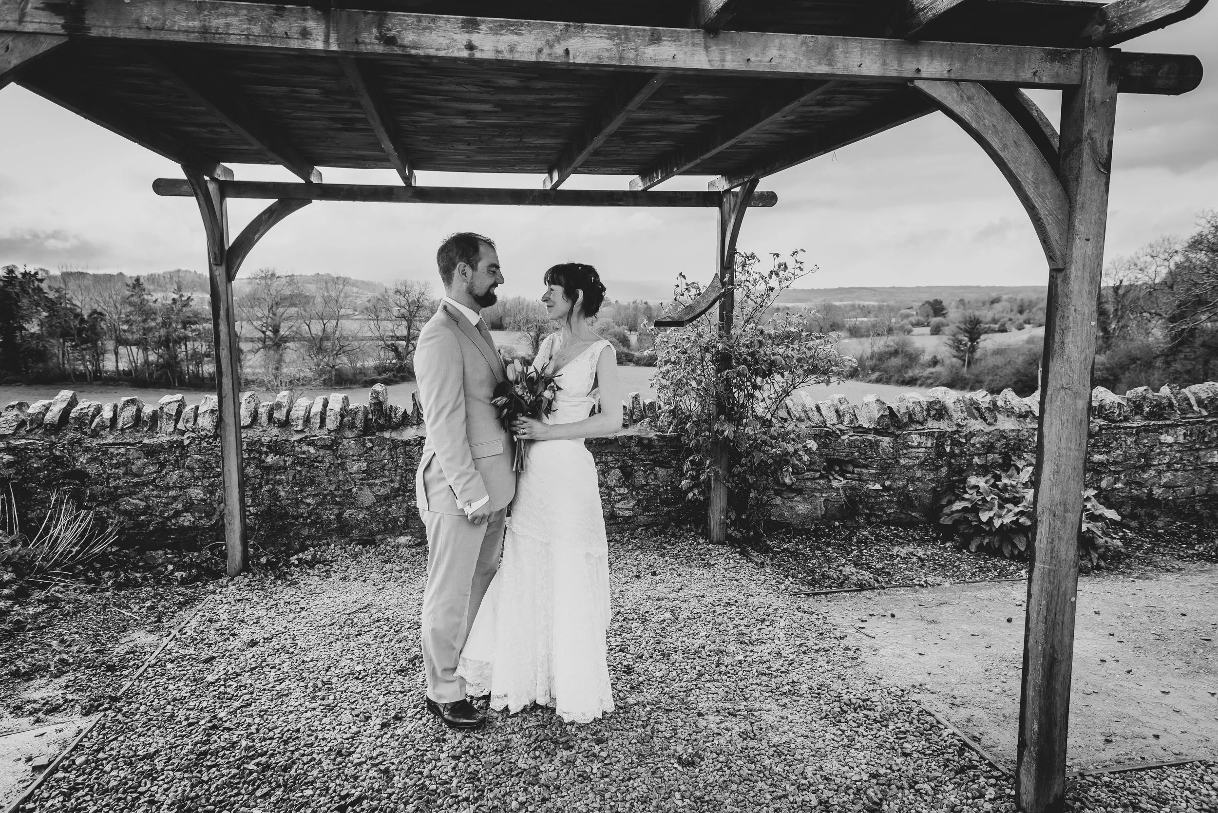 A black and white photo of a bride and groom standing under a wooden canopy outdoors, gazing into each other's eyes during their wedding ceremony with chew valley lake in background.