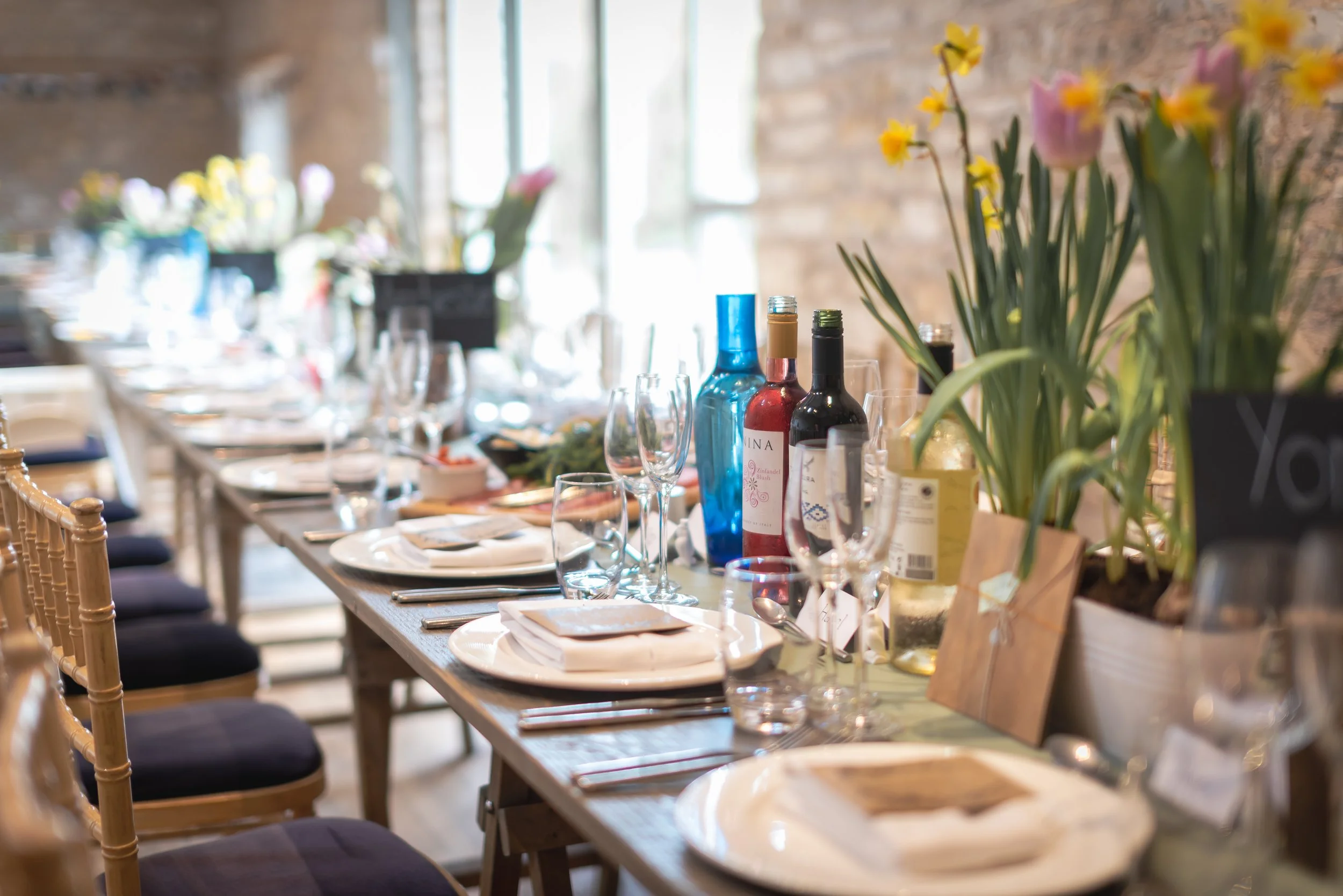 A long dining table set for a wedding breakfast with white plates, wine glasses, silverware, and folded napkins, decorated with potted plants, flowers, and bottles of wine or other beverages at Folly Farm
