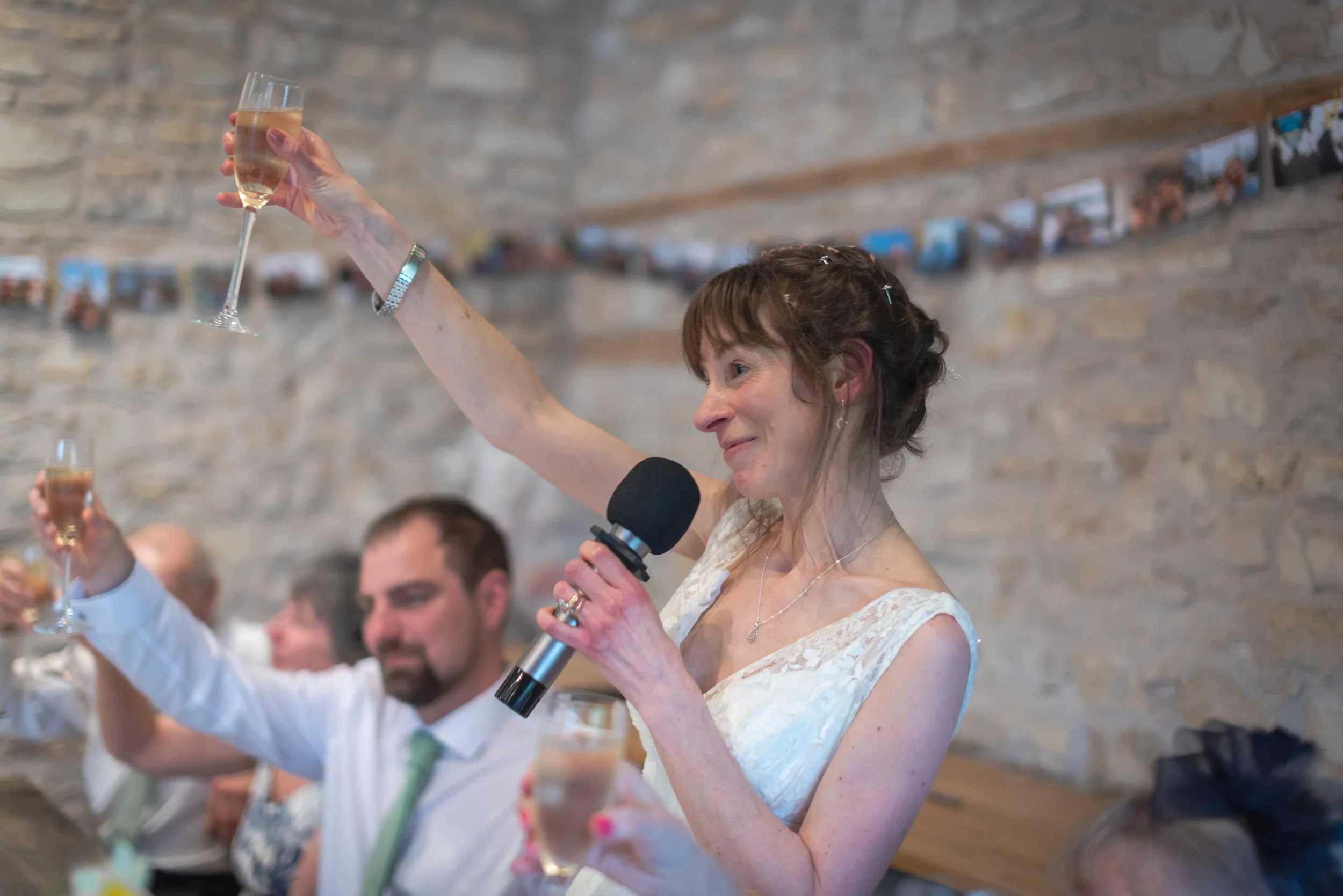 A bride in white dress giving a speech at a wedding reception, holding a microphone and raising a glass of Champagne, with guests seated at a table in the background