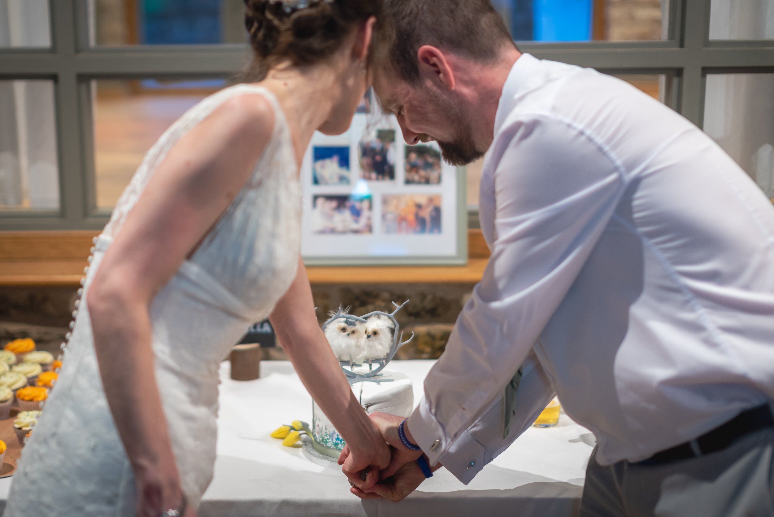 A newlywed couple holding hands and leaning close during their wedding reception, with a decorated table and photos in the background taken by South West wedding photographer