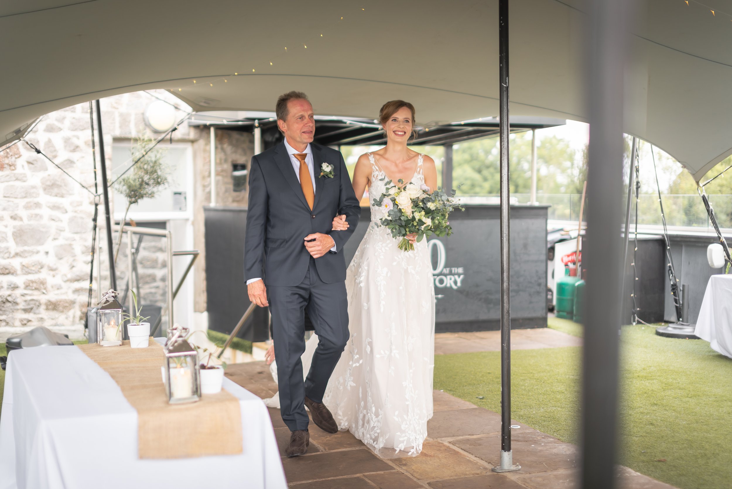 Bride walking down the aisle with her father during an outdoor wedding ceremony at Clifton Observatory in Bristol.