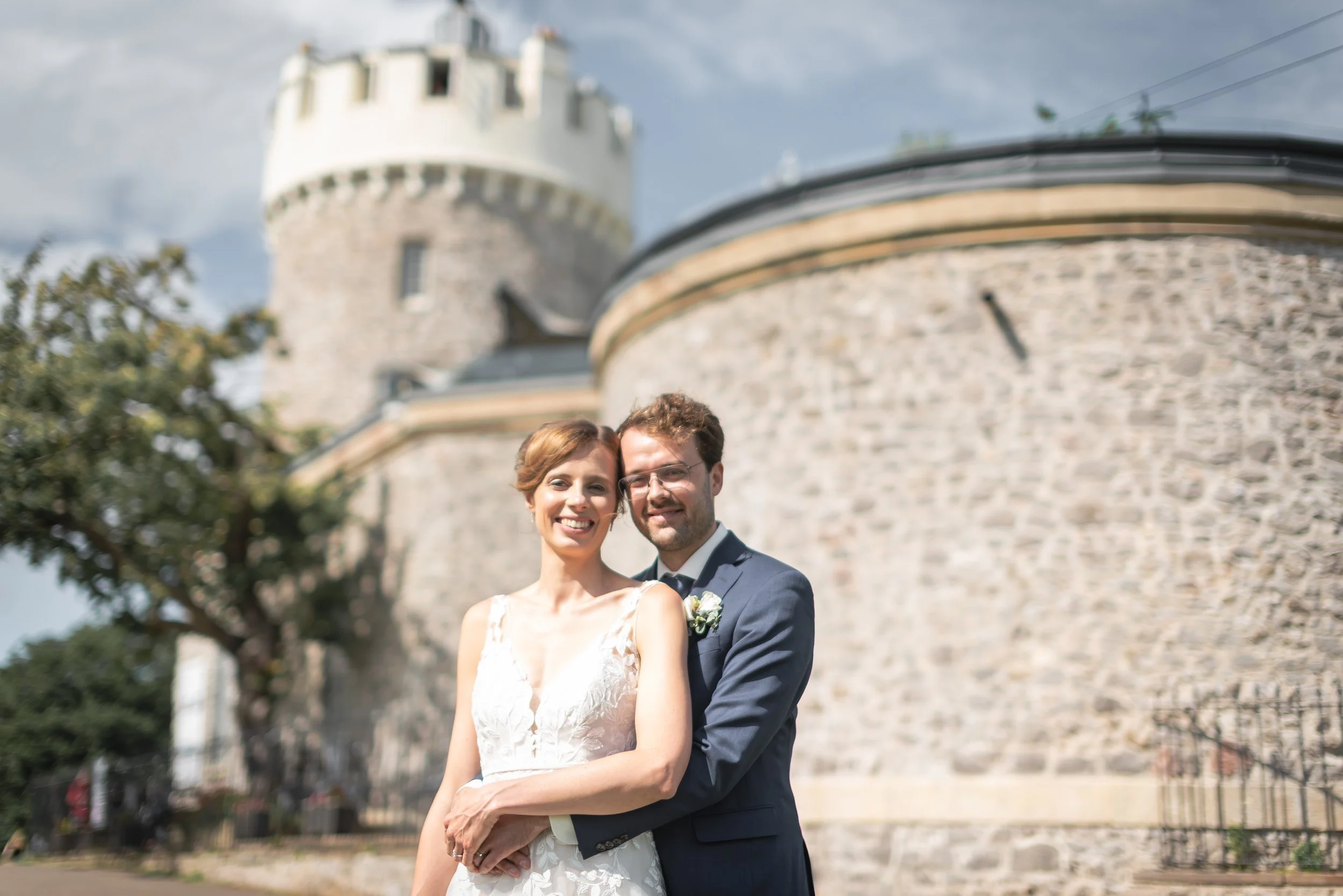 Wedding portrait of bride and groom standing in front of Clifton Observatory with panoramic views of Bristol behind them.