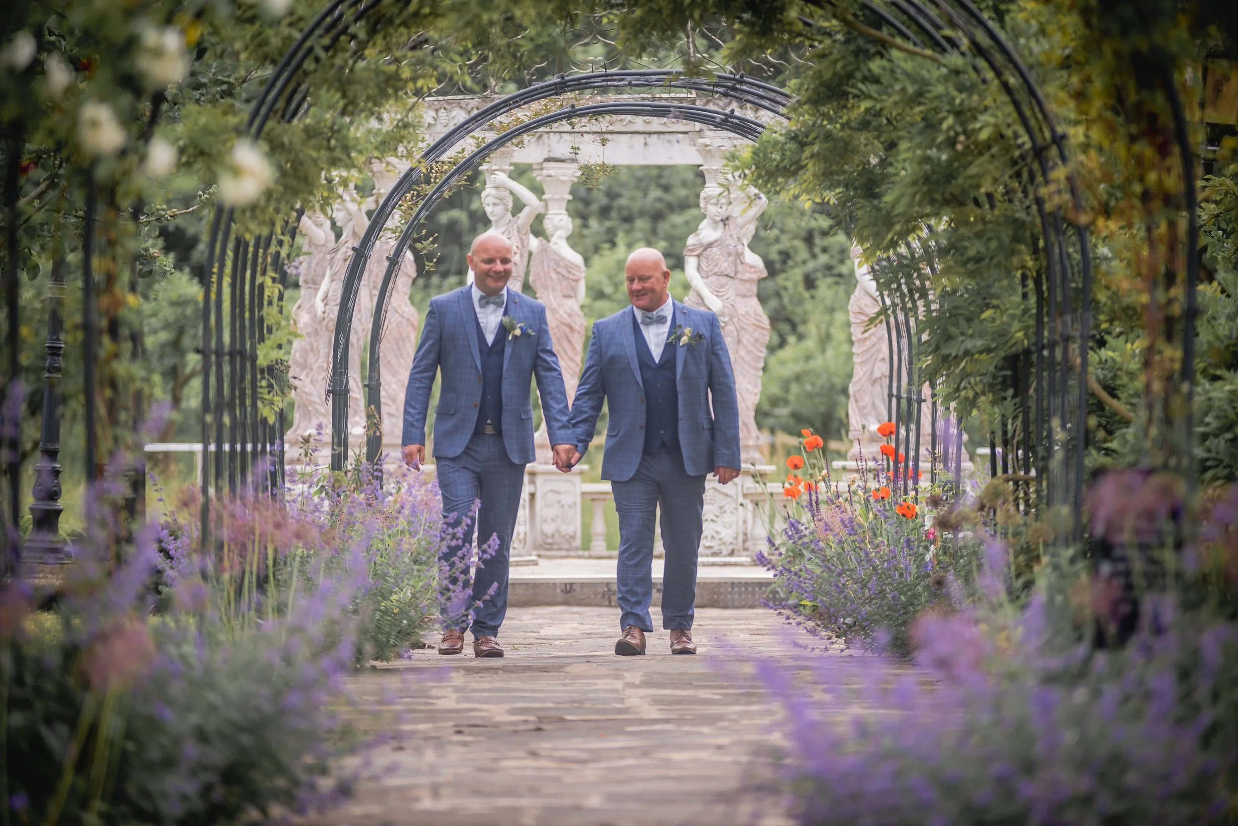 Two grooms walking hand in hand down secret garden surrounded by flowers at Berwick Lodge
