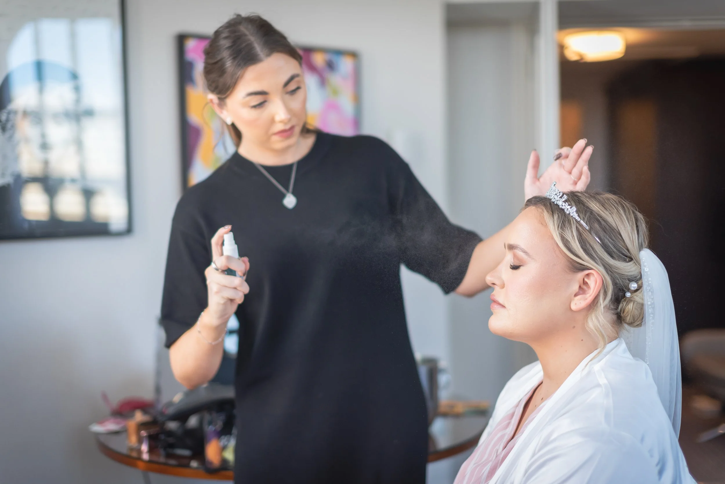Bridal makeup artist sprays finishing spray on bride's face in a well-lit room captured by South West Wedding Photographer.