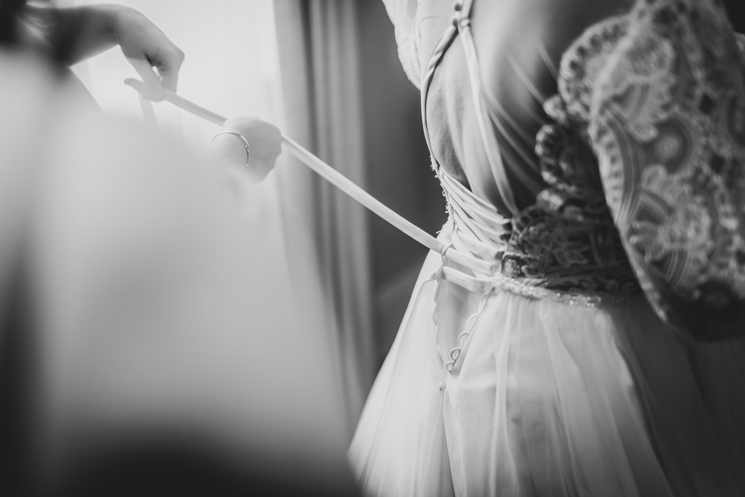 A woman in a patterned dress and a white tulle skirt is helping a bride lace up her wedding gown from behind.