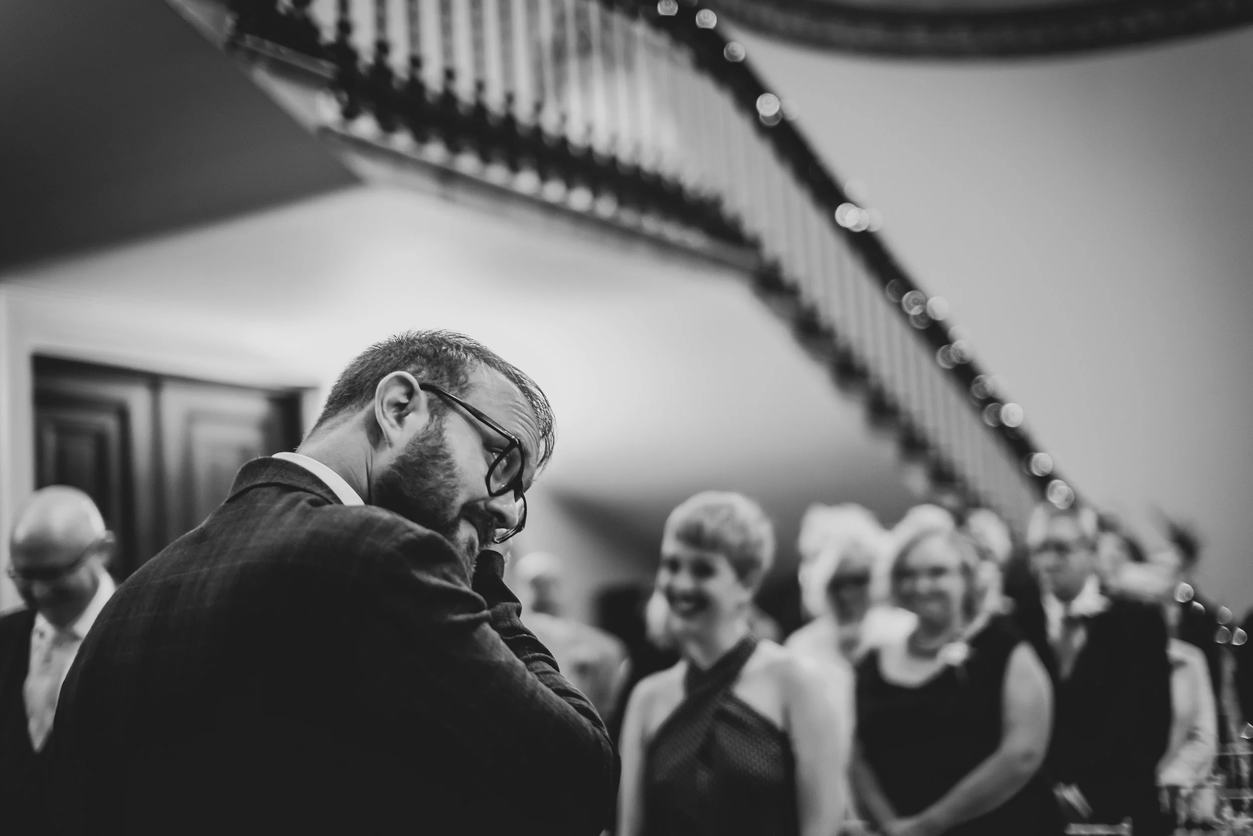 A man with glasses and a beard wearing a suit smiling and closely leaning with his head down in front of women dressed in formal attire at a social event in a room with a staircase and wood-paneled walls.