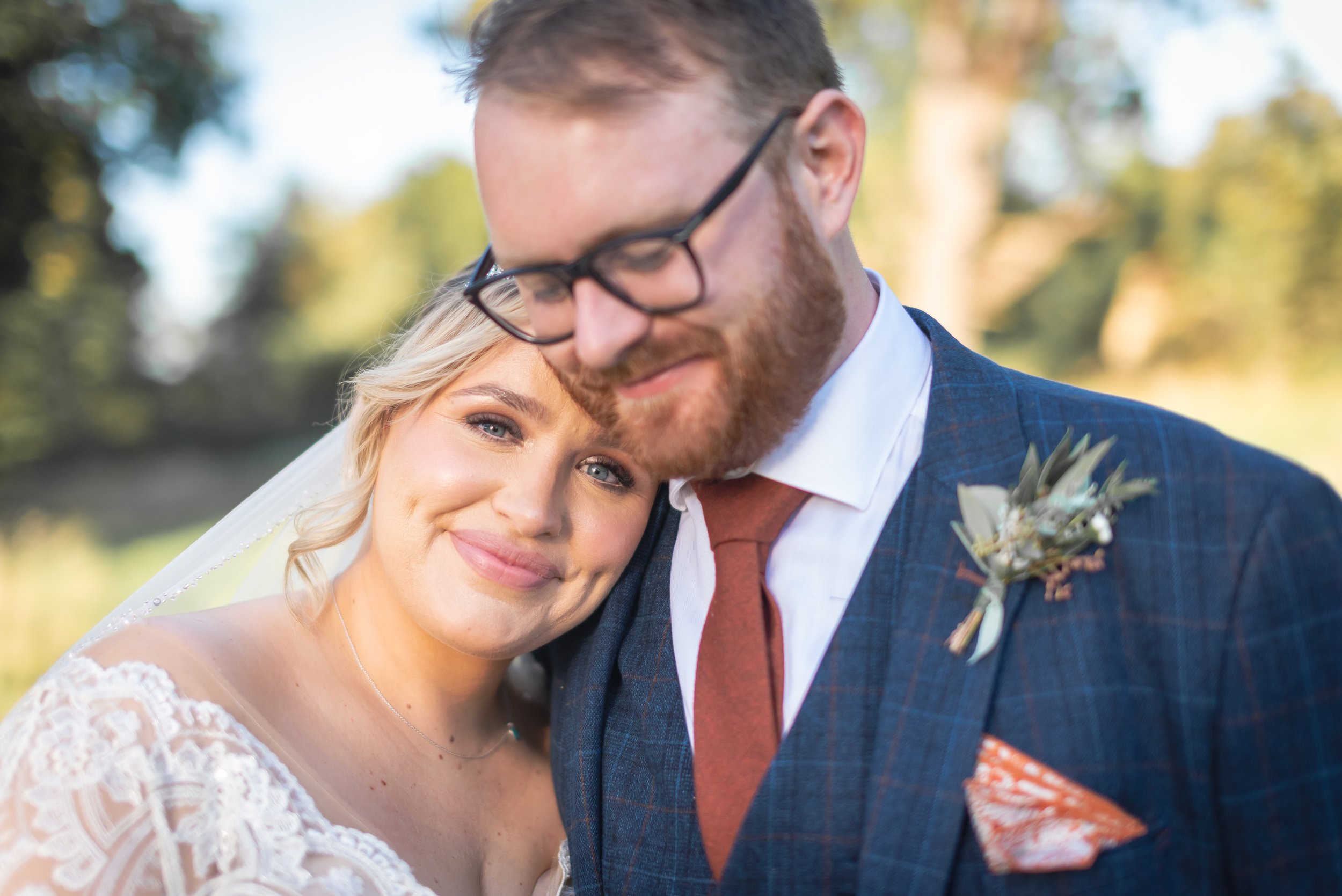 A newlywed couple in wedding attire close together outdoors, with the bride smiling softly and the groom with eyes closed, embracing each other, surrounded by blurred greenery smiling for the Bristol Wedding Photographer.