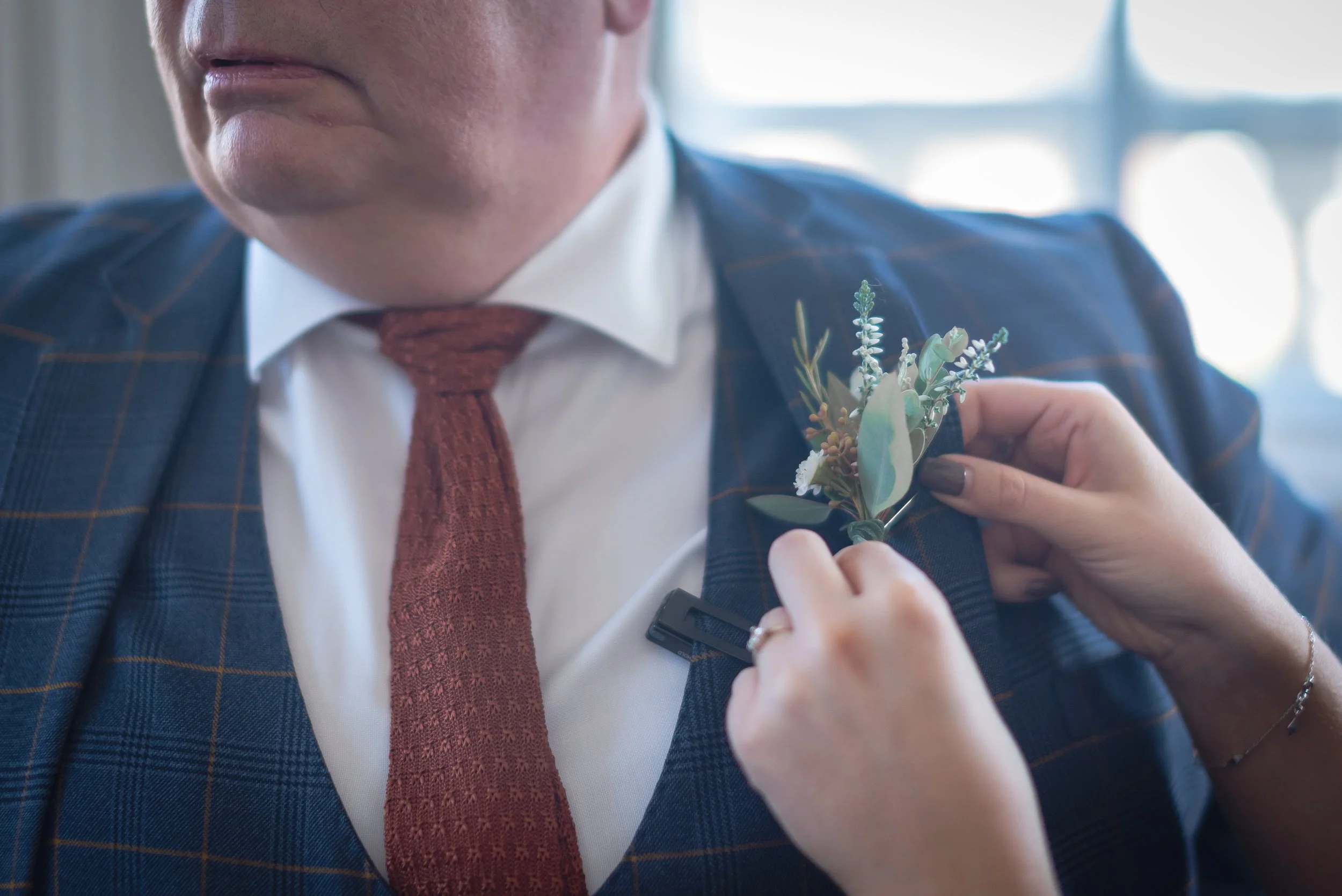 Father of the bride in a navy checkered suit with a white shirt and rust-colored tie is having a boutonniere pinned to their lapel.