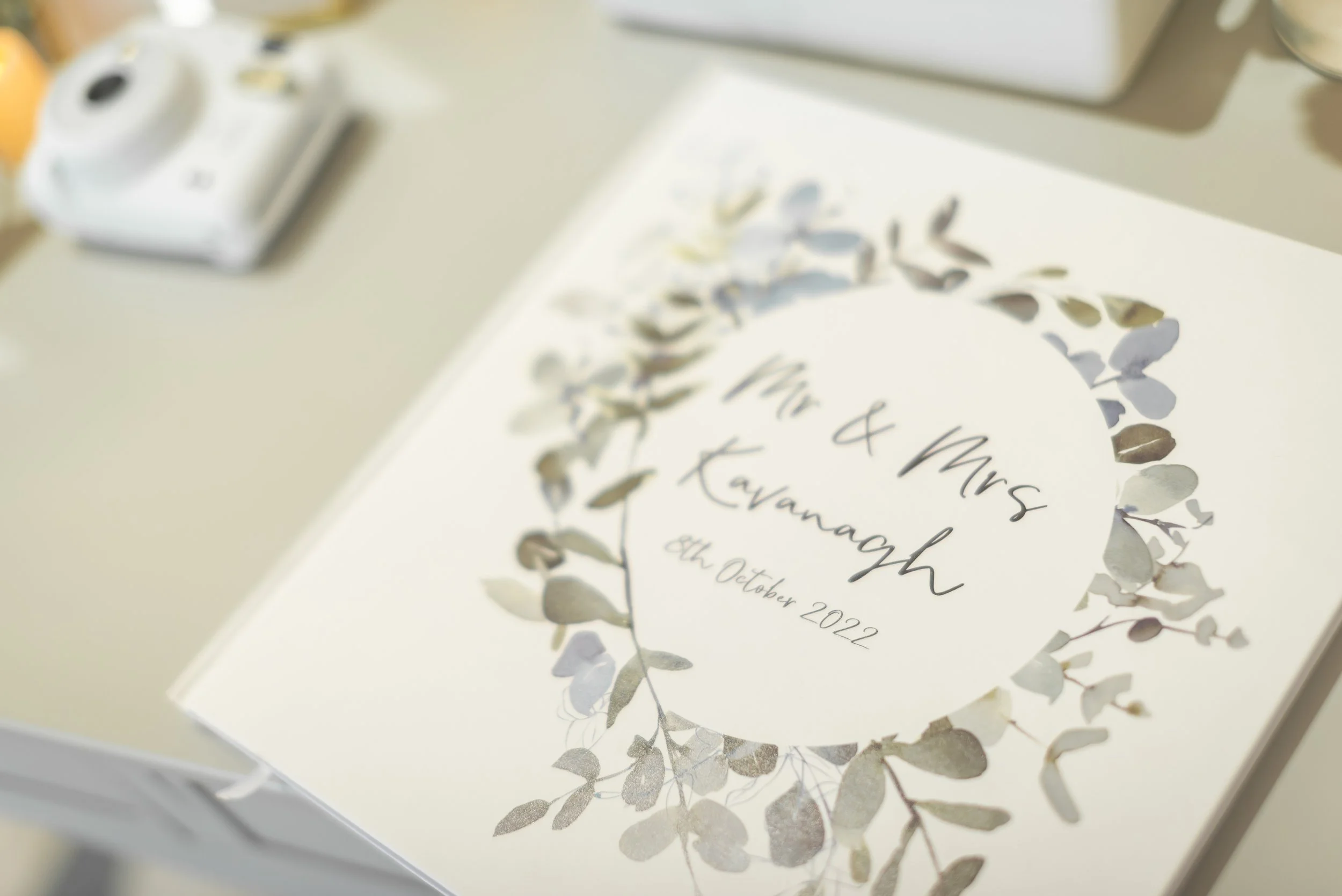 Close-up of a wedding invitation with a floral wreath, titled "Mr & Mrs. Kavanagh," dated October 8, 2022, on a white background, placed on a table with a camera and paper nearby.