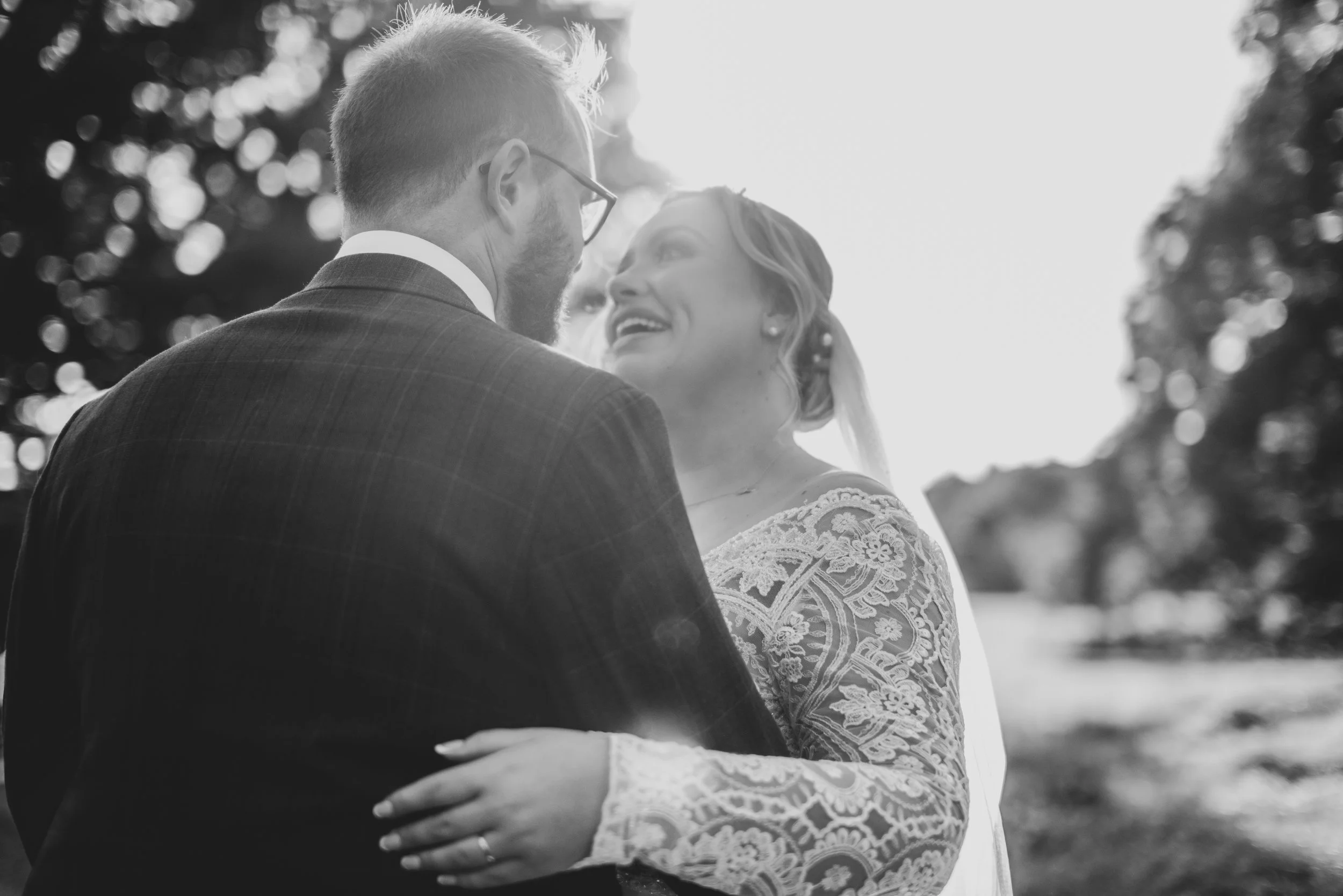 A black-and-white photo of a couple on their wedding day, embracing outdoors with trees in the background, smiling and gazing at each other.