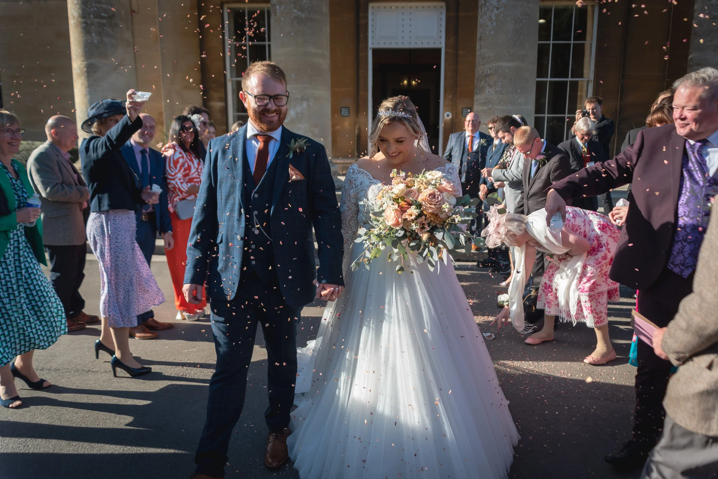 A newlywed couple walking hand in hand surrounded by friends and family outside a building, celebrating their wedding with confetti at Leigh Court near Bristol