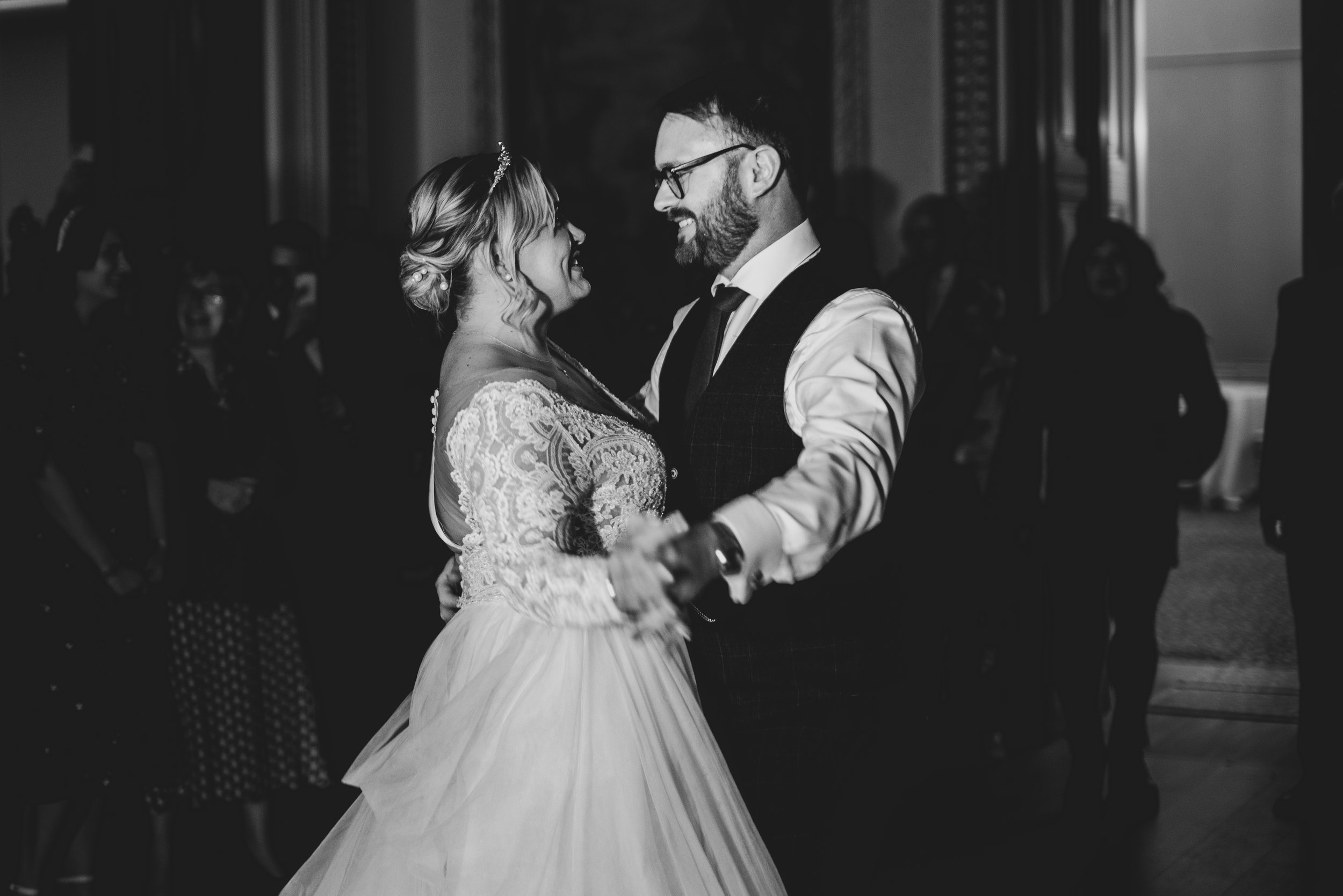 A black and white photo of a bride and groom dancing together at a wedding reception, surrounded by guests during the first dance in the ballroom at Leigh Court.