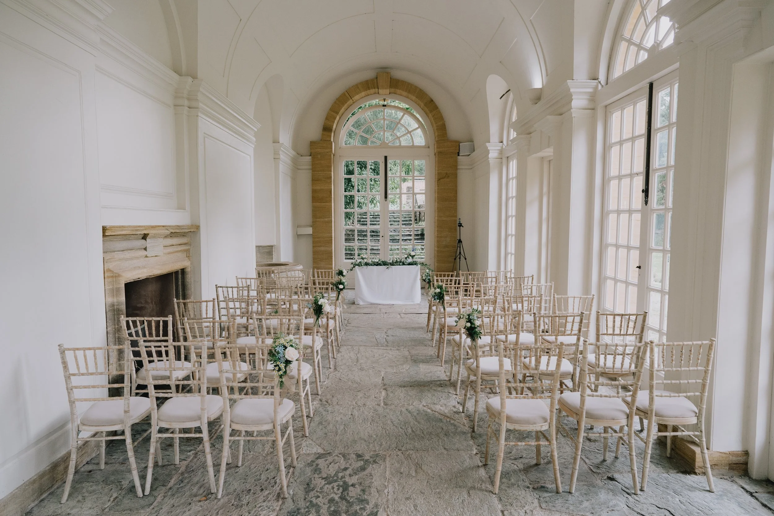 Wedding ceremony setup in a bright, elegant room with arched windows and white walls, featuring rows of wooden chairs decorated with small floral arrangements, facing an altar with a window backdrop at Hestercombe Gardens