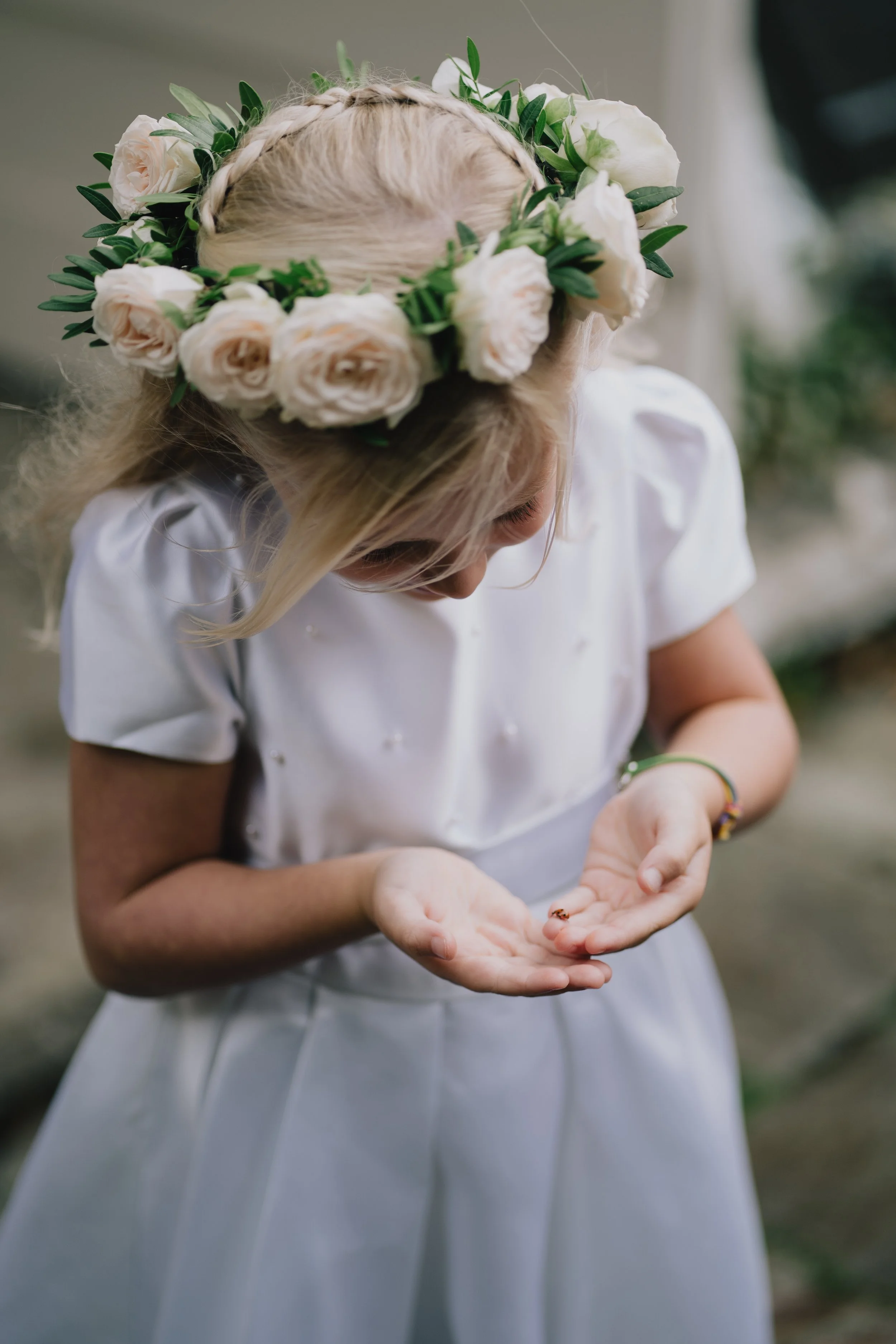 A young girl with blonde hair adorned with a floral crown of white roses and greenery, looking at and holding a ladybug on her hand, dressed in a white dress taken by Canid Wedding Photographer