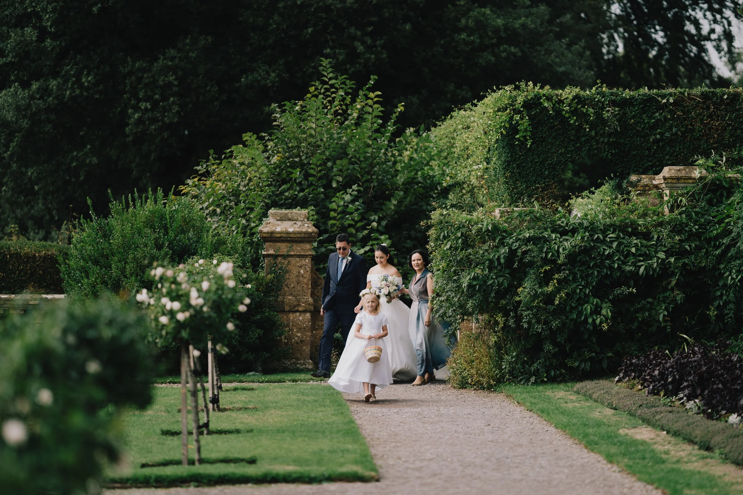 A wedding procession with three women and one man walking on a garden path surrounded by greenery and bushes ready for wedding at Hestercombe Gardens.