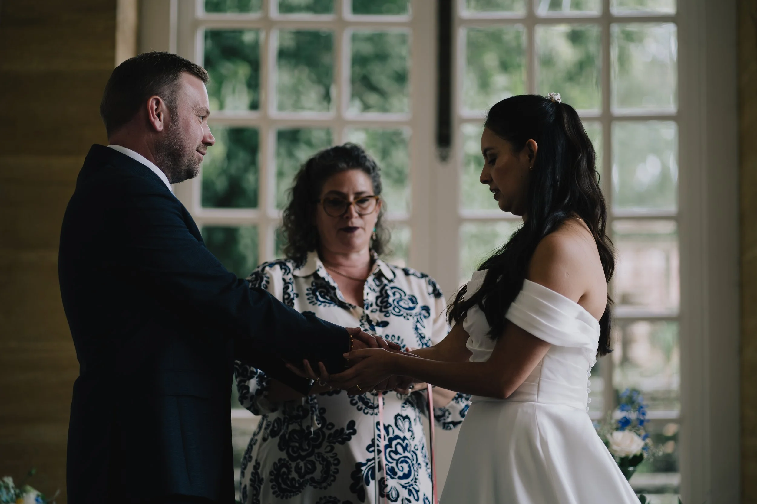 A couple exchanging wedding vows in a ceremony, with a woman officiant standing behind them, inside a room with large windows and greenery outside, Hestercombe Gardens.