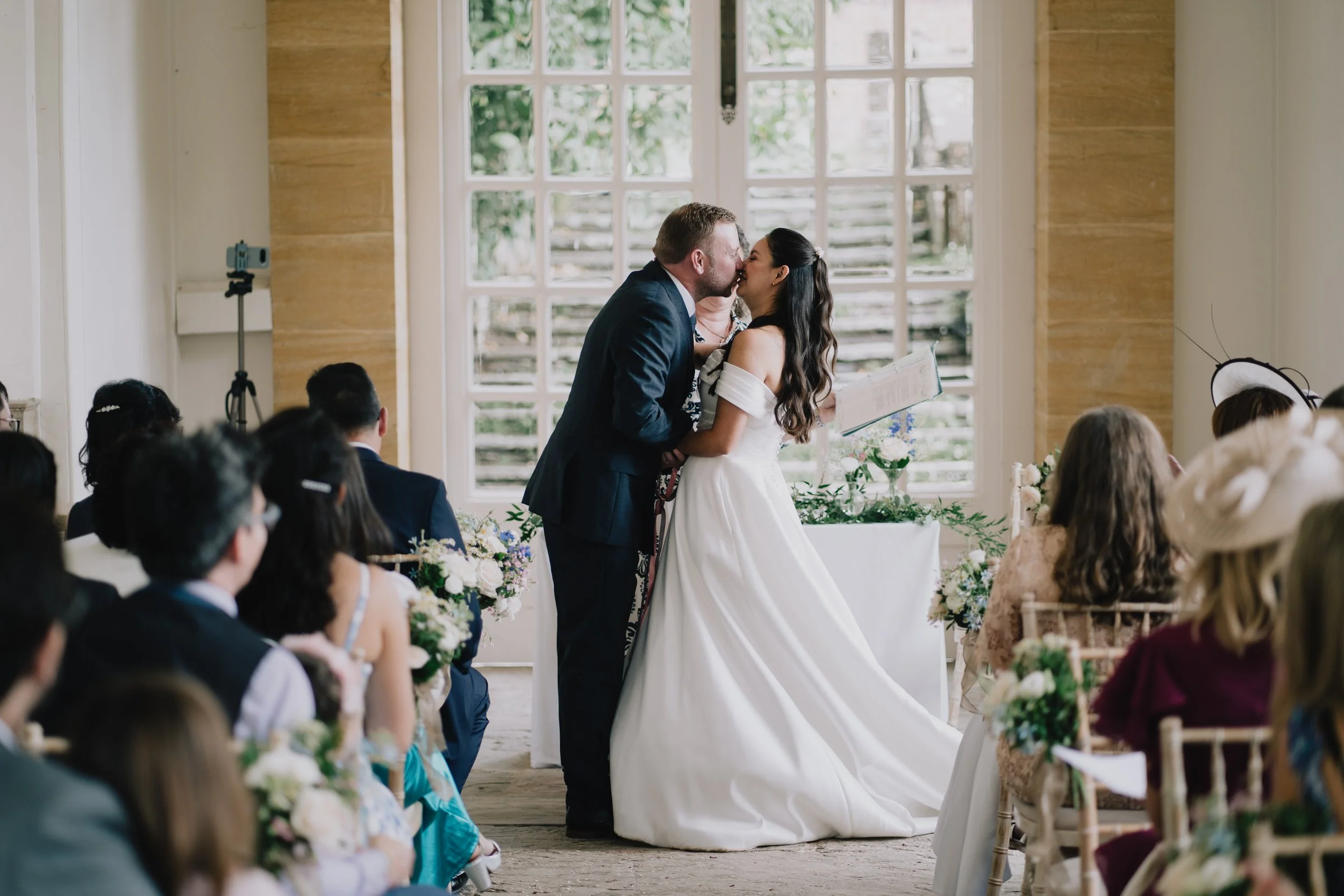 A bride and groom kiss during their wedding ceremony, surrounded by seated guests in a sunlit Hestercombe Gardens.