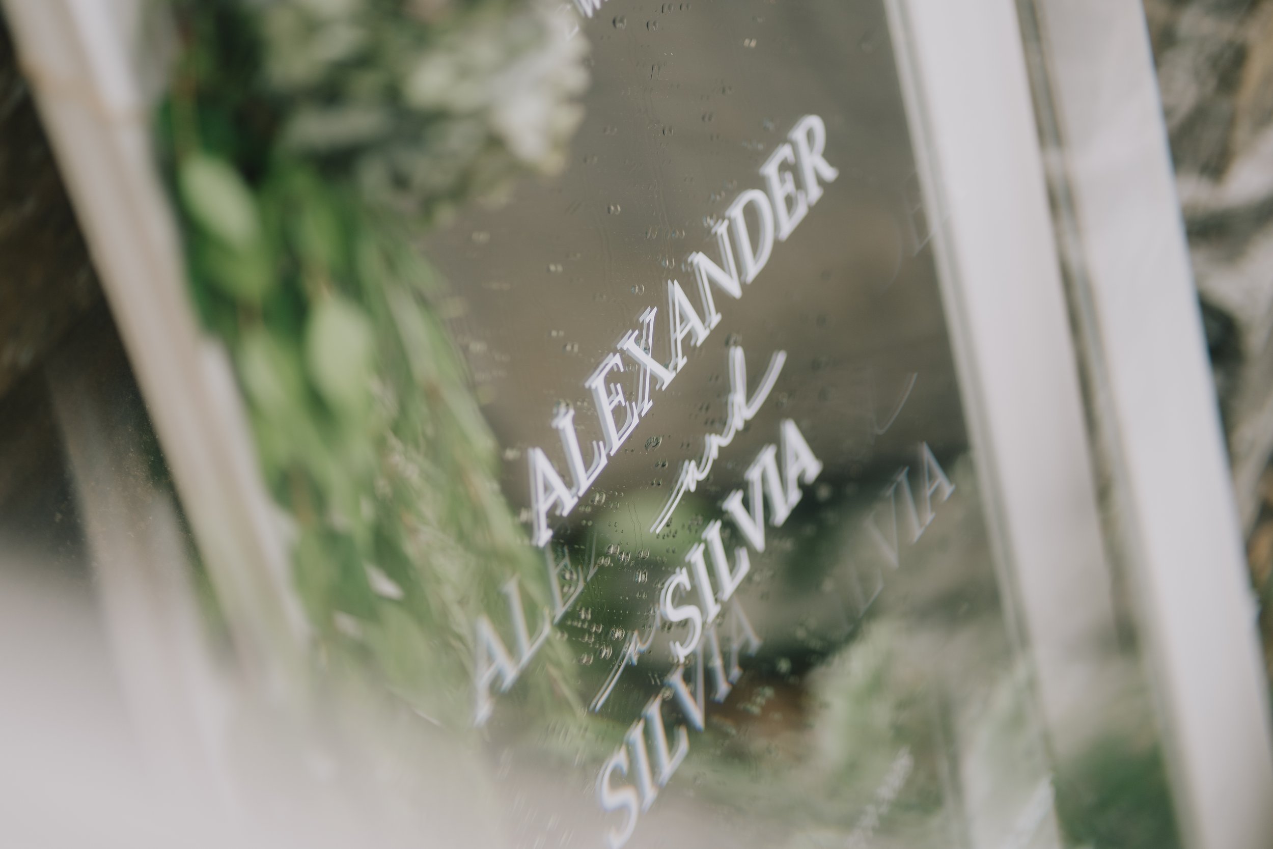 A reflective metallic surface with the engraved text "ALEXANDER the SILVIA". The surface has water droplets on it, and there are blurred green leaves in the background taken by relaxed Bristol Wedding Photographer.