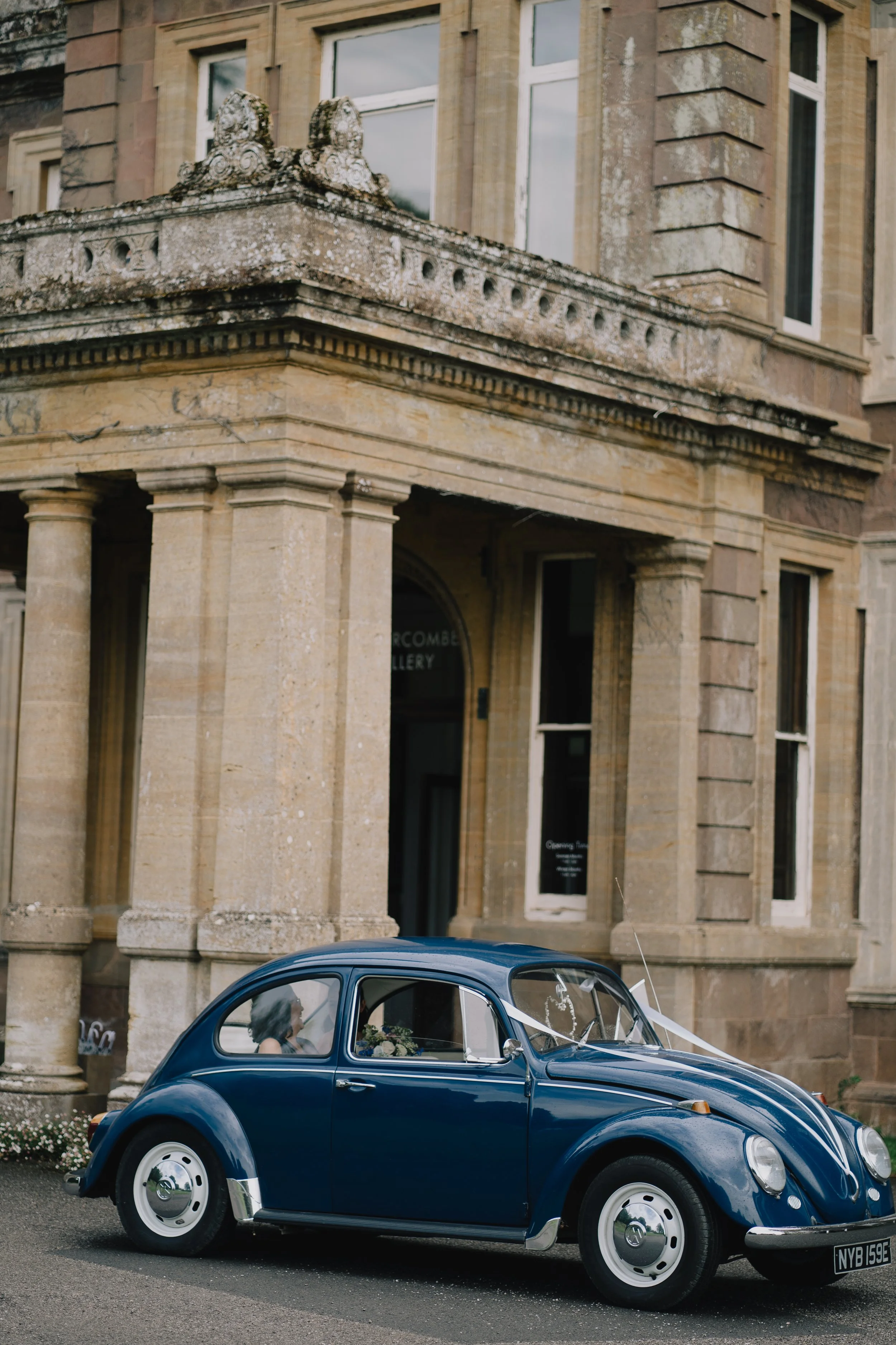 A vintage blue Volkswagen Beetle parked in front of a historic stone Hestercombe Gardens with large windows and decorative architectural details.