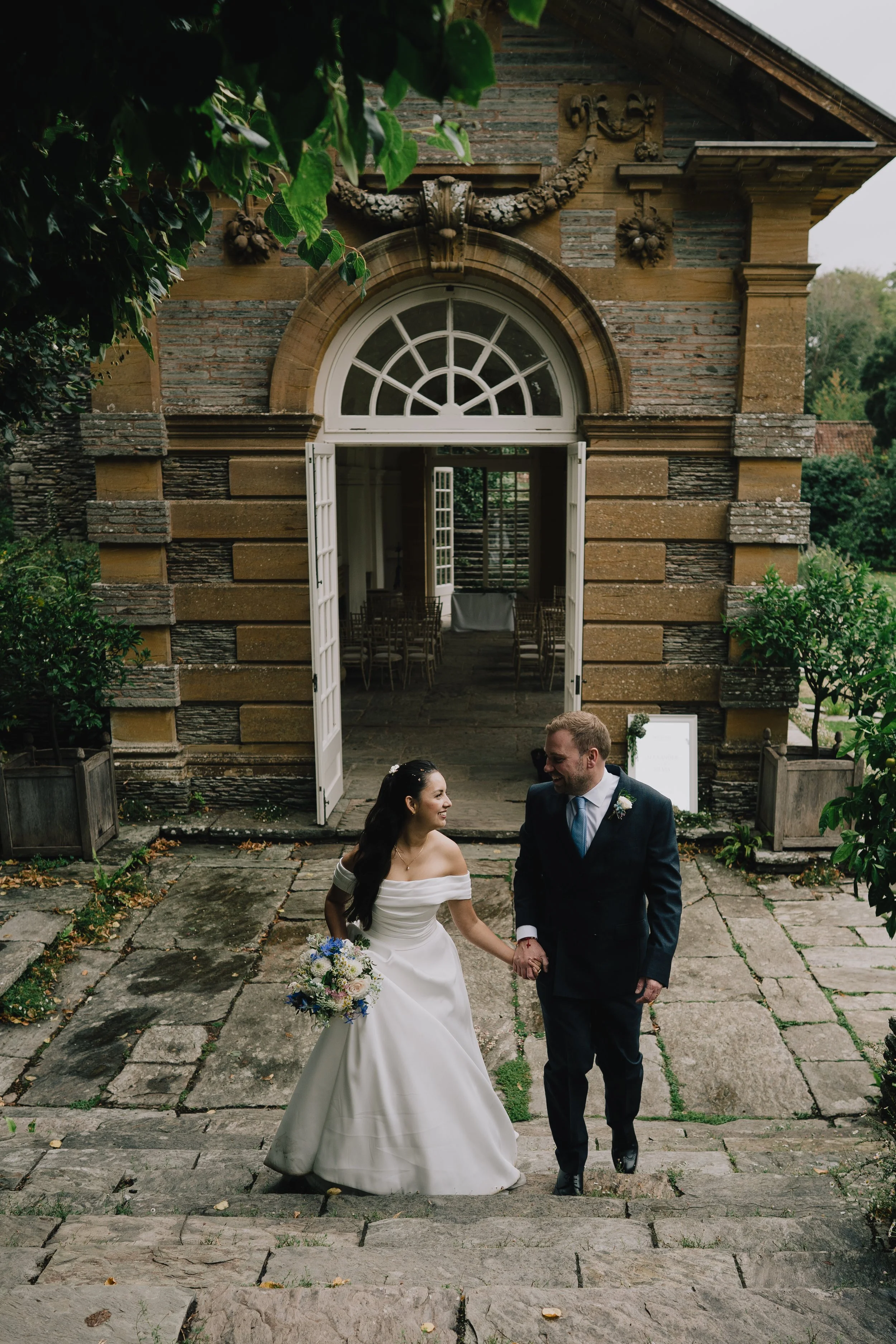 A bride in a white wedding dress and a groom in a dark suit holding hands and smiling while walking down stone steps outside a rustic stone building with open white double doors after tieing the knot at Hestercombe Gardens.