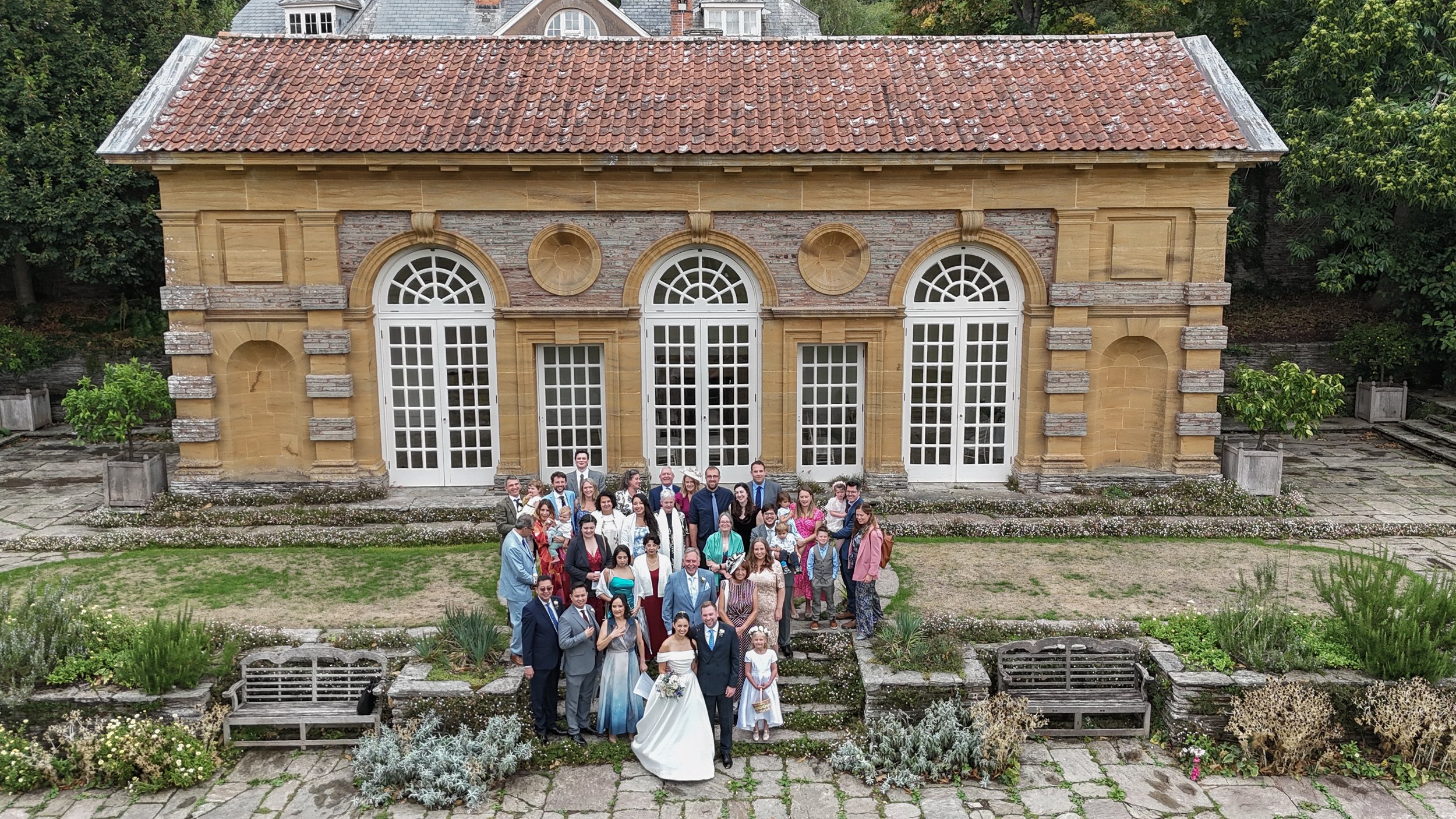 Group of people dressed in formal attire standing on steps in front of a historic stone building with large white arched windows and patio doors, surrounded by a garden taken by drone at Hestercombe Gardens near Taunton