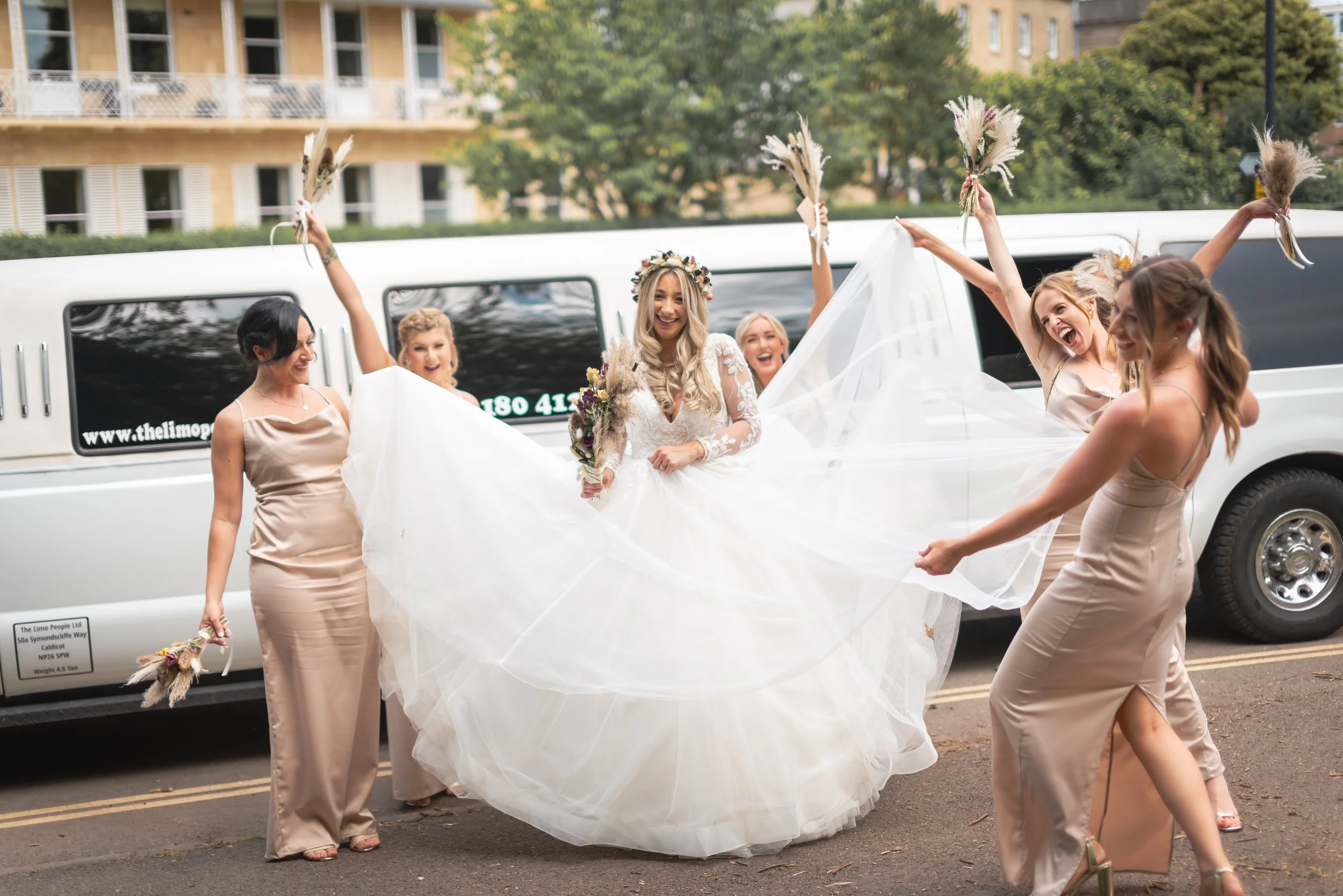 Bride in a white wedding gown holding a bouquet, surrounded by bridesmaids in beige dresses playing with her veil, outside in front of a limo ready for her wedding a Clifton Observatory in Bristol
