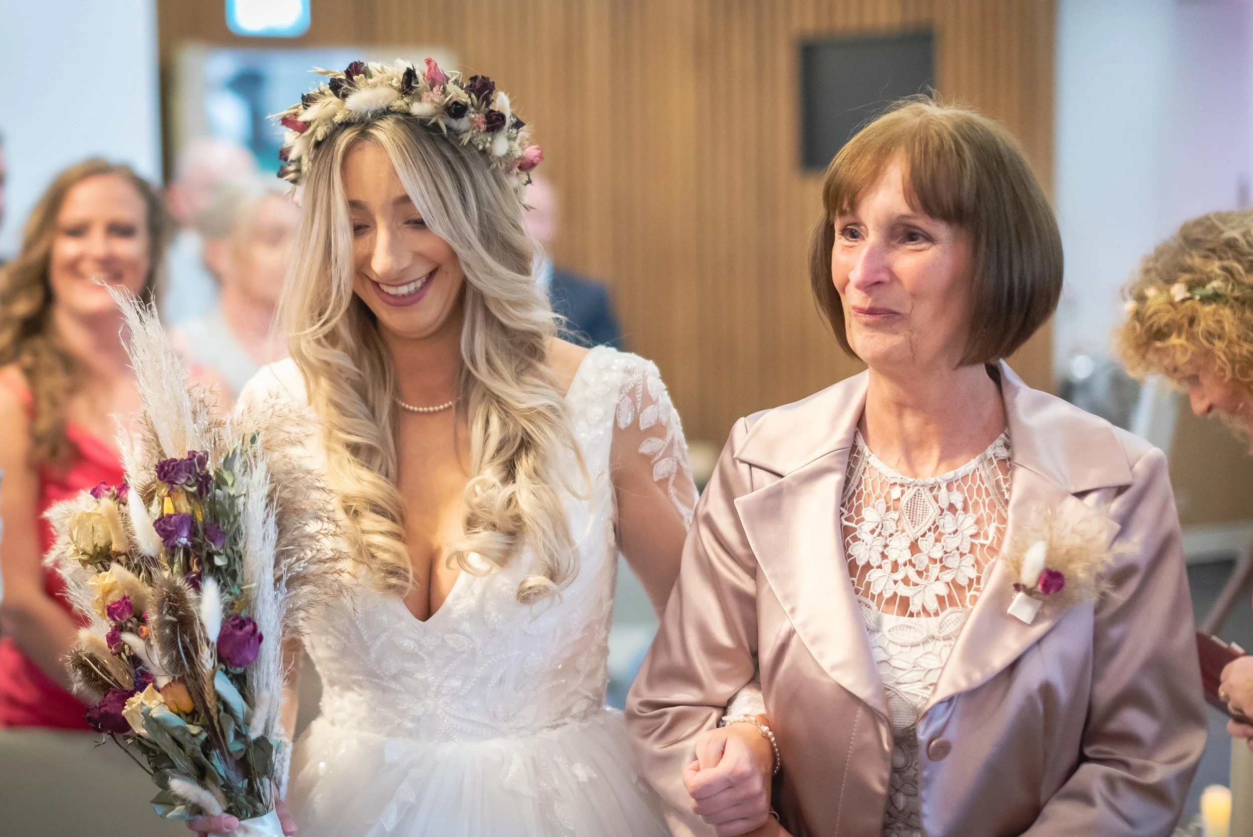 A bride in a white wedding dress with a floral crown, holding a bouquet, smiling as she walks arm-in-arm with an older woman in a light-colored blazer, at a wedding ceremony.
