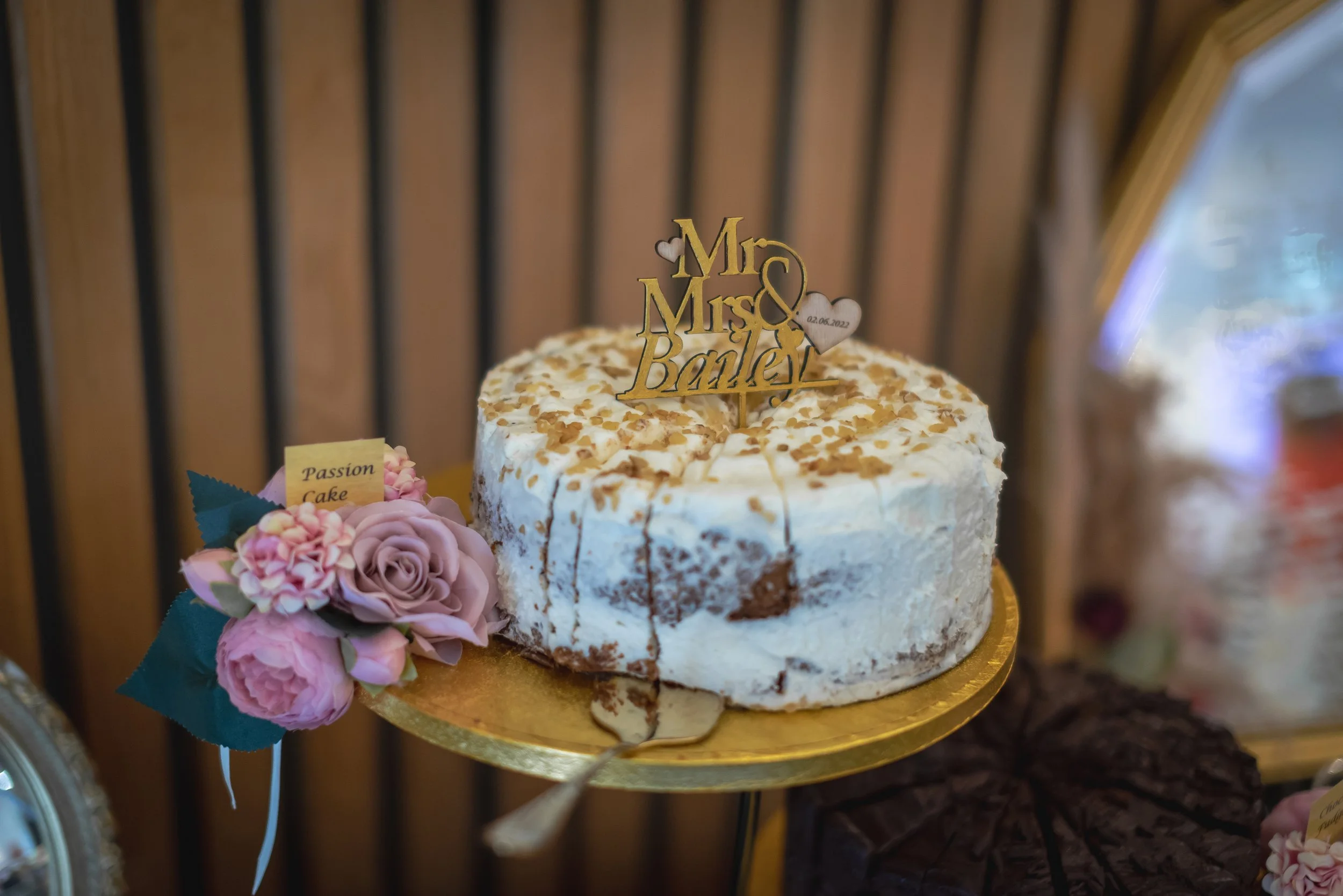 Cake with a "Mr & Mrs Bailey" topper, surrounded by flowers, on a golden cake stand.