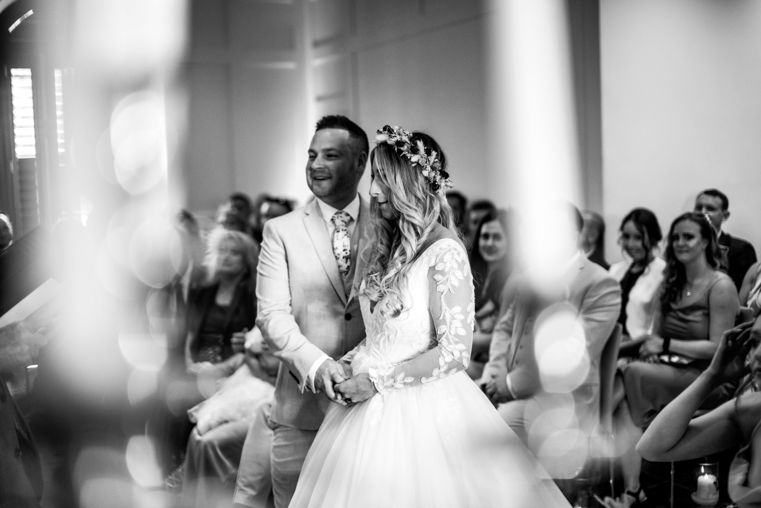 Black and white photo of a wedding ceremony with a bride and groom holding hands, surrounded by seated guests.