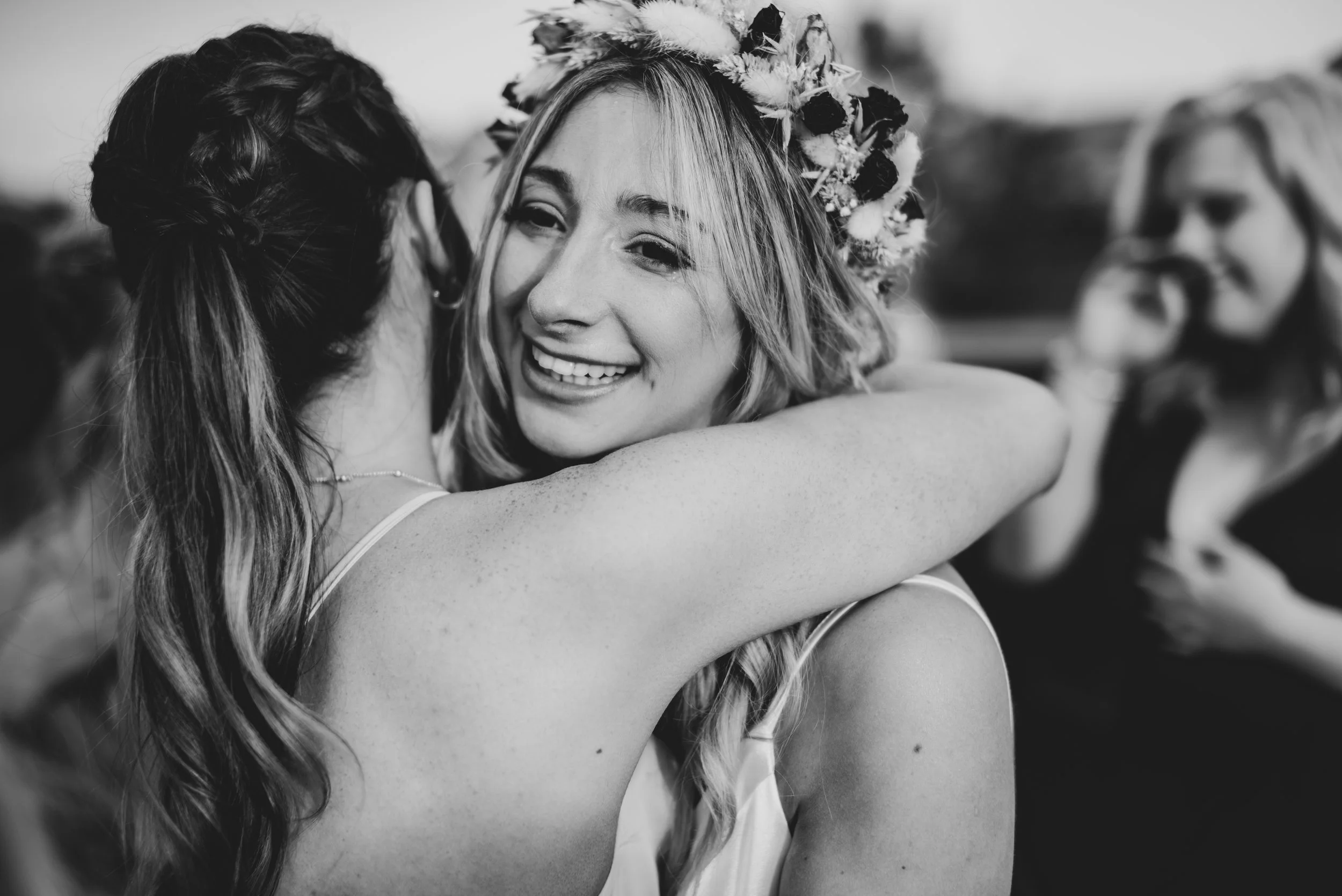Two women hugging, one smiling with a flower crown, in a joyful moment at a celebration, with a third woman in the background.