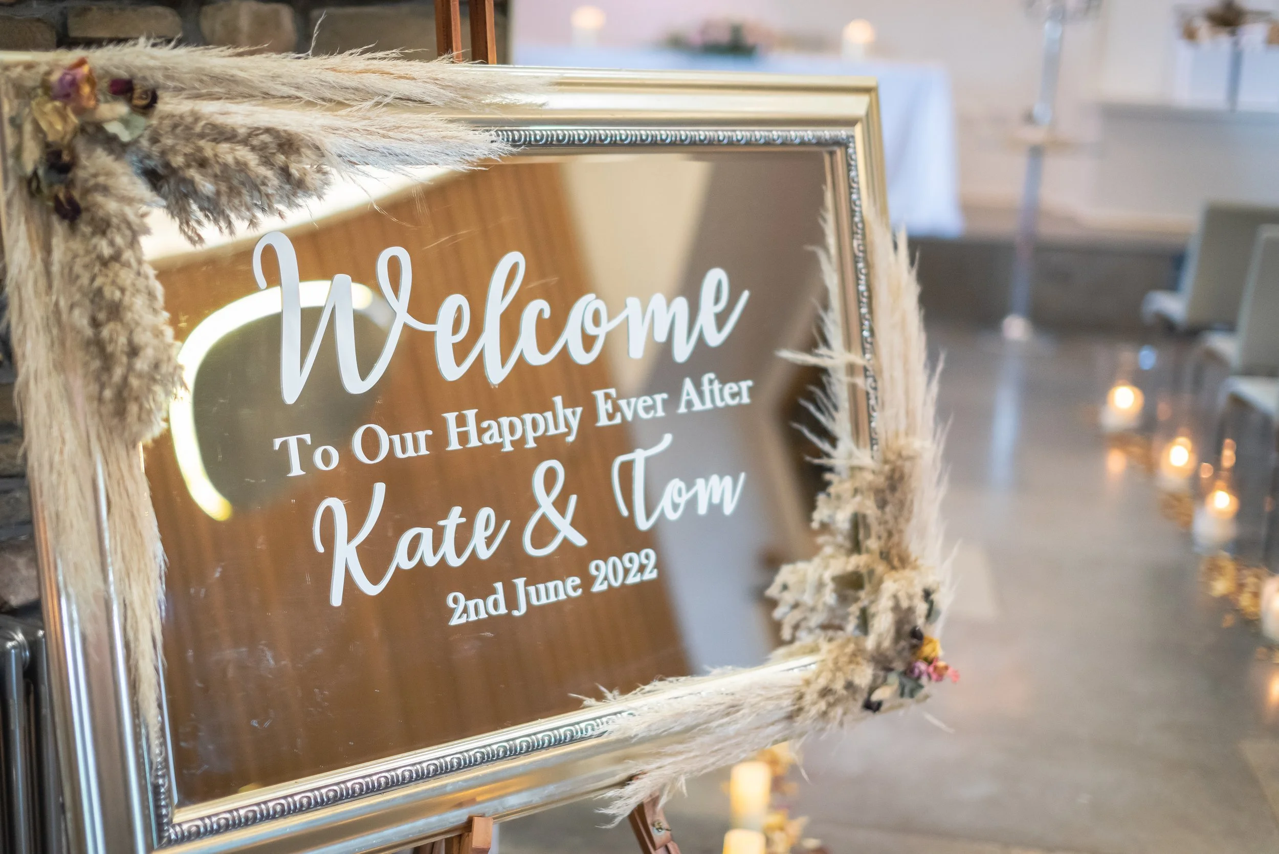 Wedding welcome sign with floral decorations, reading 'Welcome To Our Happily Ever After Kate & Tom 2nd June 2022', in a decorated indoor venue.