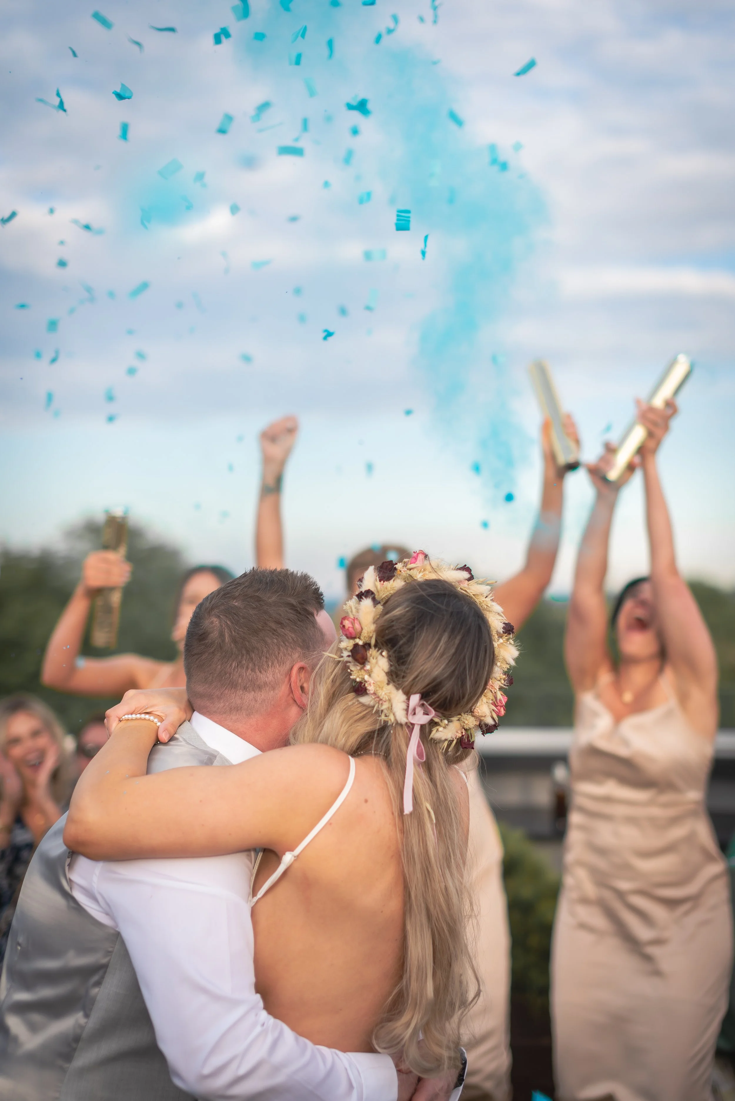 A newlywed couple hugging each other while wedding guests celebrate with confetti and party blowers in the background during a gender reveal at their wedding