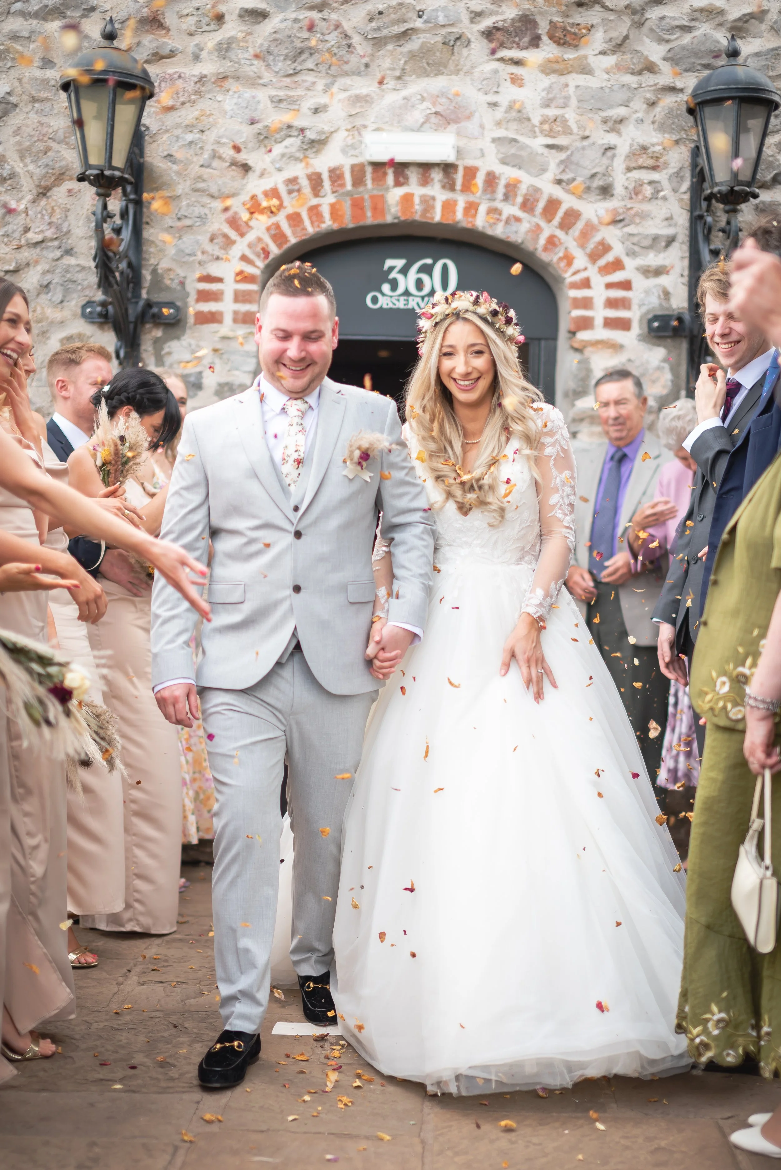 Happy newlywed couple holding hands and smiling as they walk through a shower of flower petals, surrounded by friends and family outside Clifton Observatory following their wedding