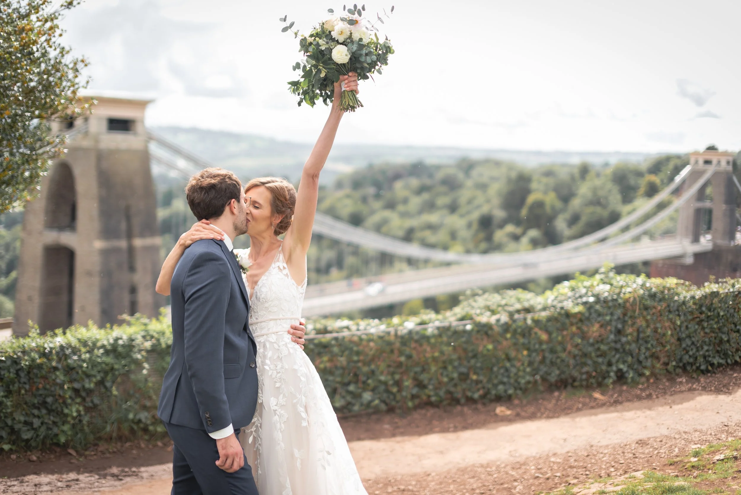 A bride and groom sharing a kiss outdoors, with the bride holding a bouquet of flowers above their heads. The background features a the Clifton Suspension Bridge in Bristol.