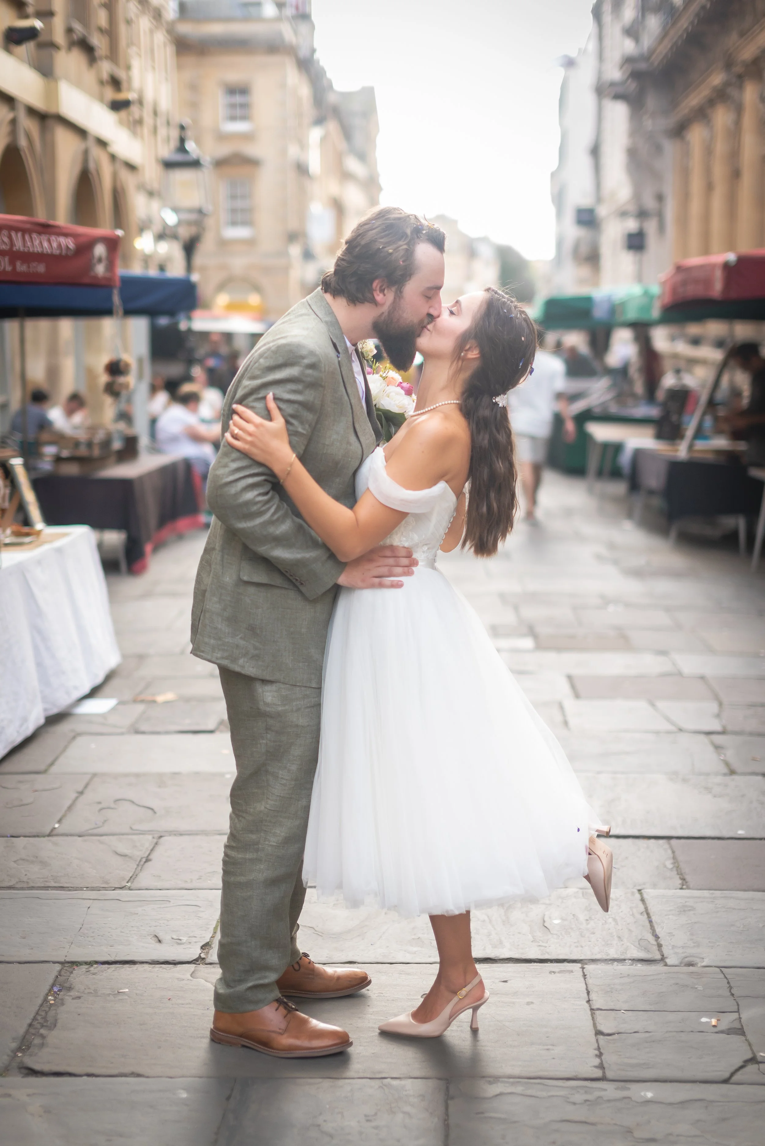 A romantic couple sharing a kiss on a historic city street, with outdoor market stalls and old buildings on Corn Street in Bristol.