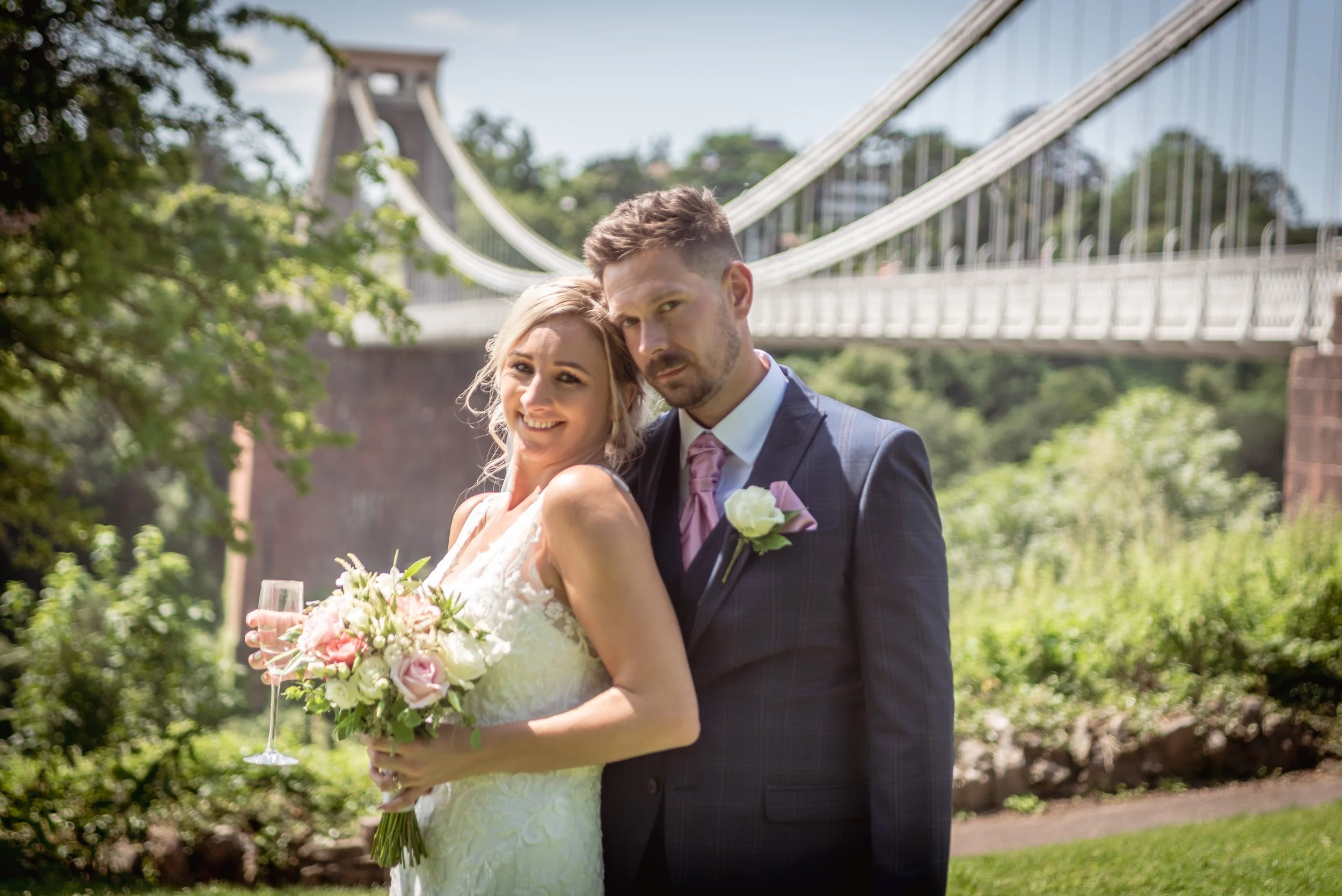A newlywed couple standing outdoors with the suspension bridge in the background. The bride is holding a bouquet and a glass of champagne, smiling at the camera. The groom is dressed in a dark suit with a pink tie and a white boutonniere, looking serious.