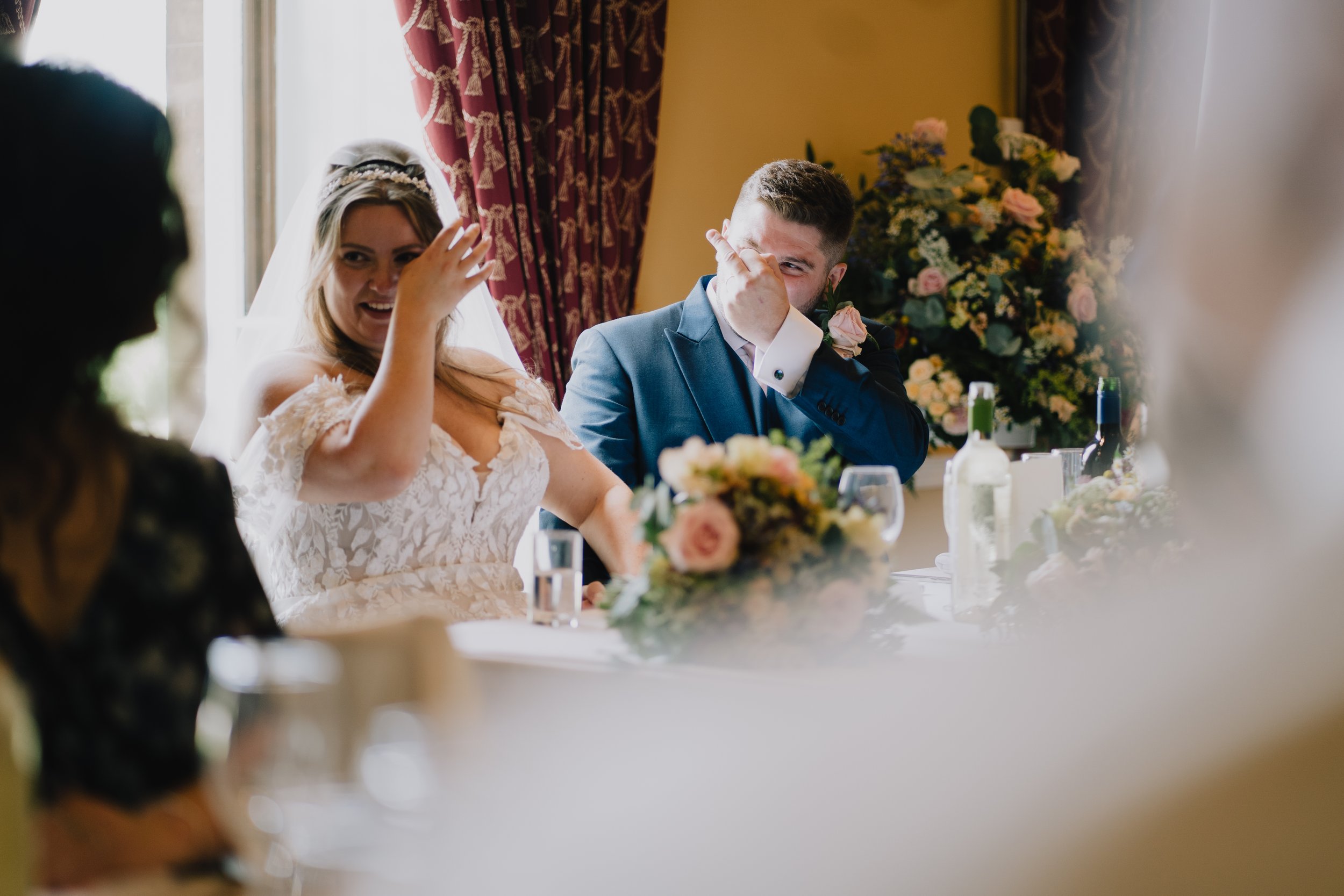 A bride and groom sitting at a wedding reception table, smiling and laughing, with guests partially visible around them, floral arrangements, and a window with curtains in the background at Leigh Court.