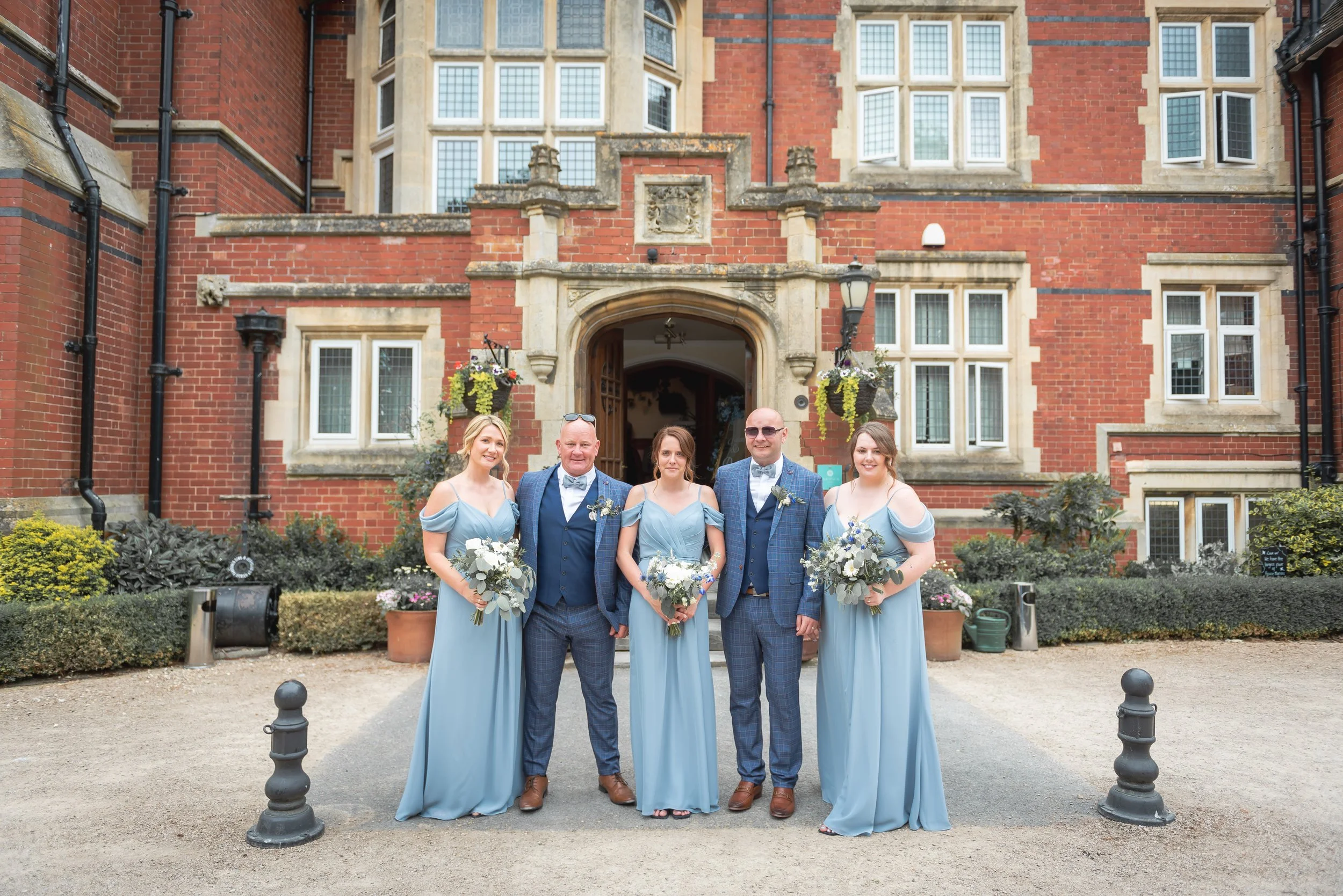 Group of five people dressed for a wedding, standing in front of a historic red brick building with multiple windows and decorative stone archway entrance at Berwick Lodge.