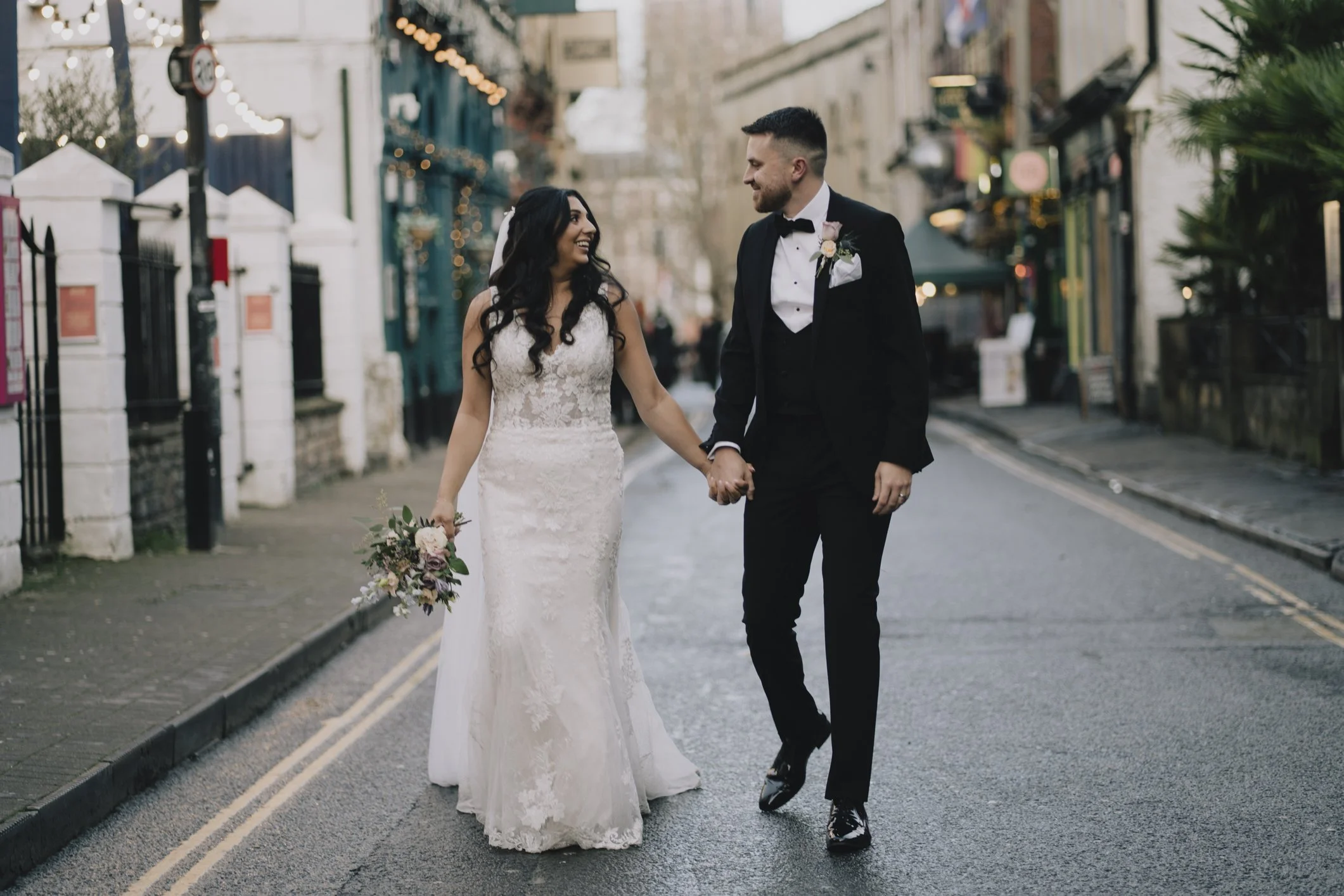 A newlywed couple holding hands and smiling at each other while walking on a city street decorated with string lights and buildings, the bride in a lace wedding dress holding a bouquet, and the groom in a black tuxedo in Bristol.