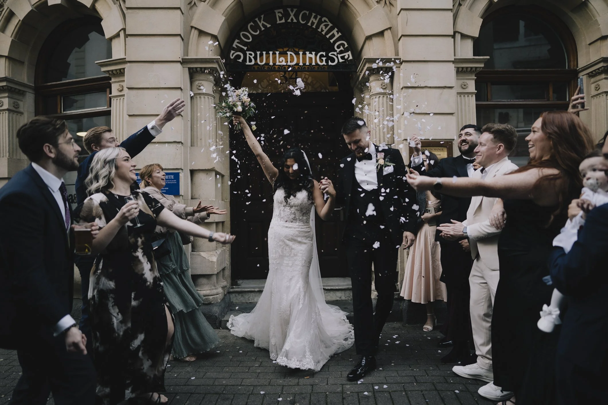 A bride and groom celebrating outside a building labeled 'Stock Exchange Buildings' as friends and family throw confetti around them taken by Bristol Wedding Photographer