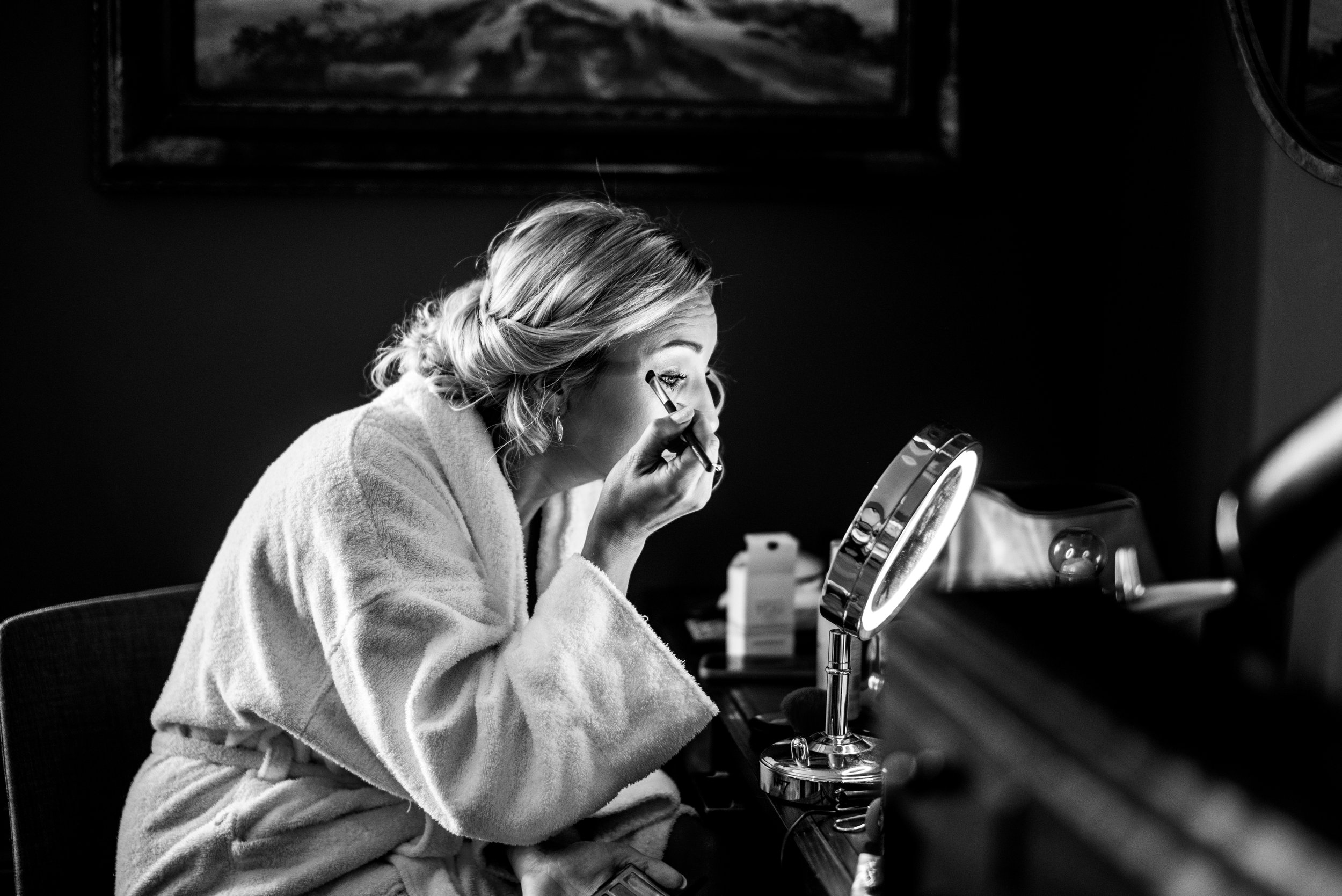 A woman sitting at a vanity table applying makeup in front of a round mirror with built-in light, wearing a fluffy robe, with a framed painting hanging on the wall behind her in Clifton near Bristol.