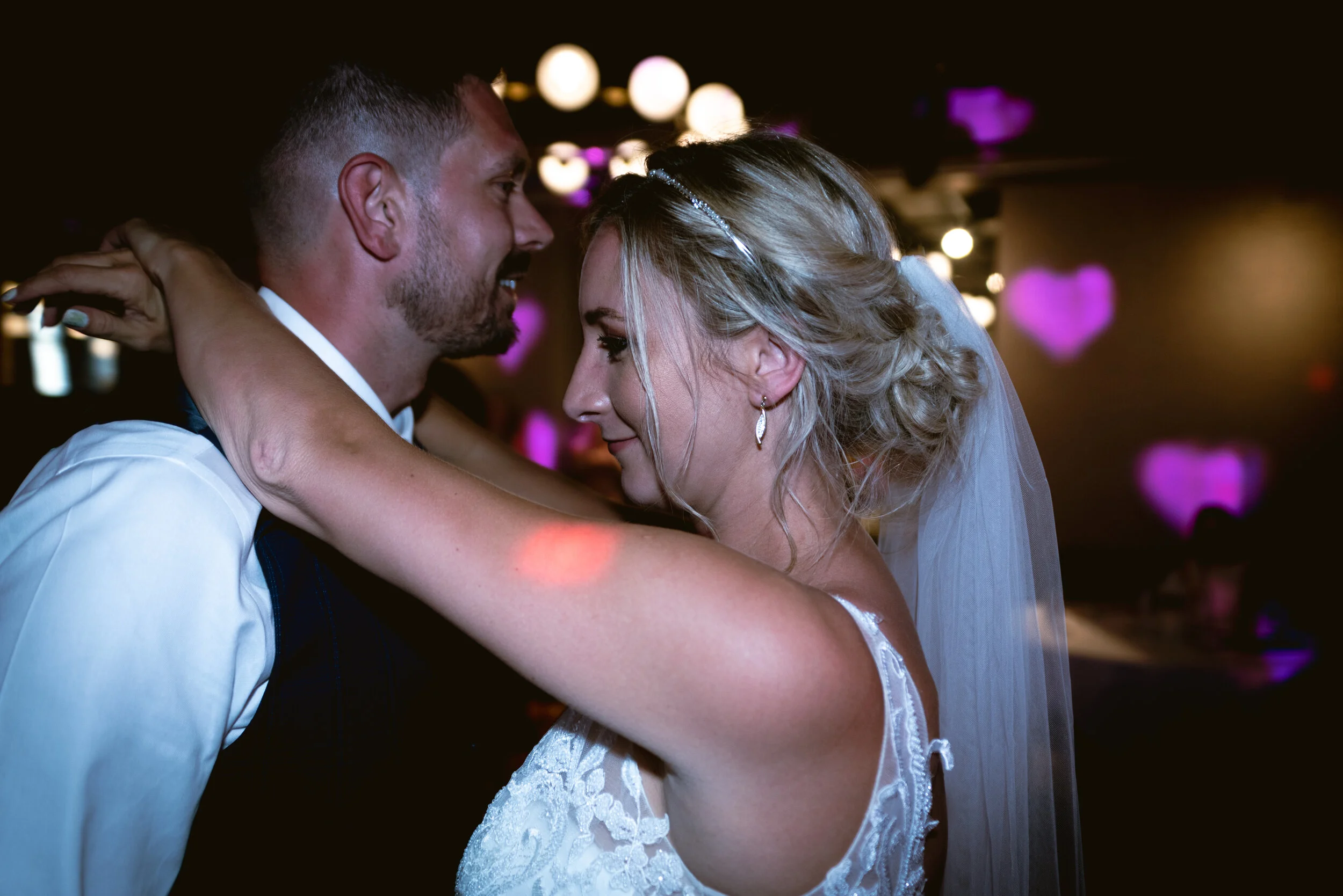 A bride and groom dancing closely during their wedding reception, smiling with their faces close together, with heart-shaped purple lights in the background captured by Avon Gorge Hotel Wedding Photographer