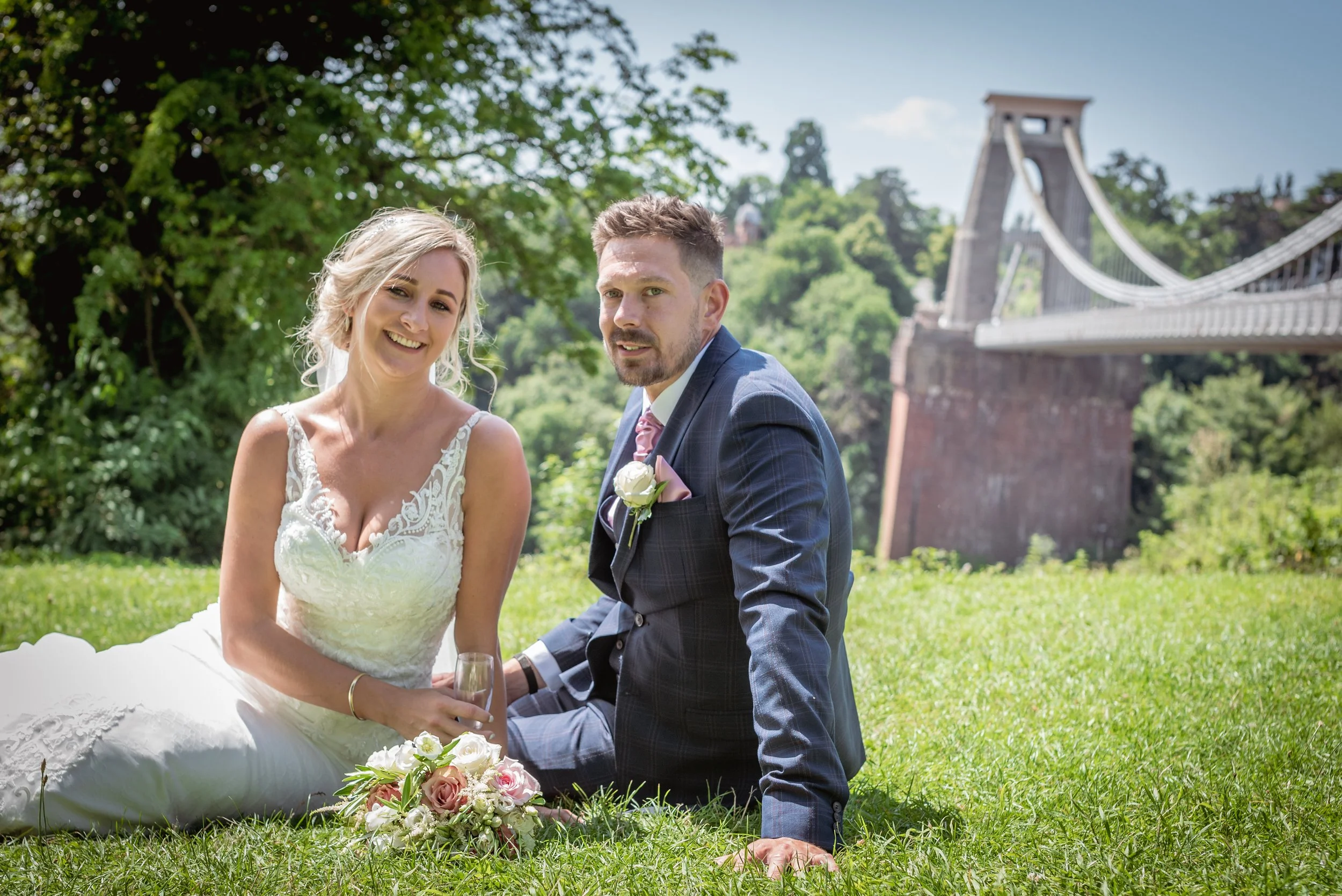 A bride and groom sitting on the grass near Clifton Suspension Bridge, smiling and holding a glass of champagne, with a wedding bouquet in front of them.