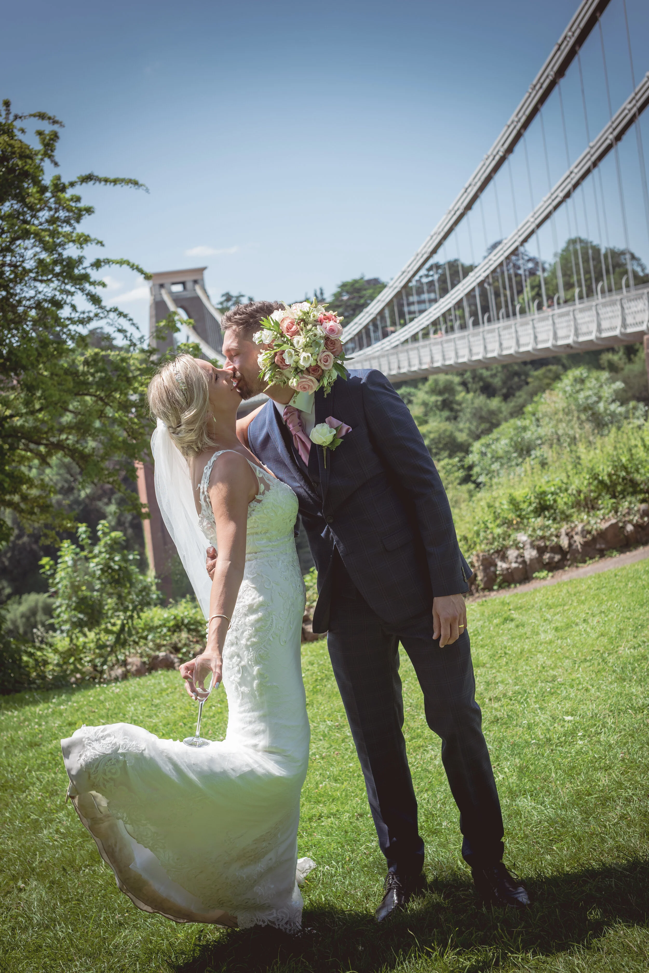A bride and groom sharing a kiss outdoors under Clifton Suspension Bridge on a sunny day, with green grass and trees in the background.