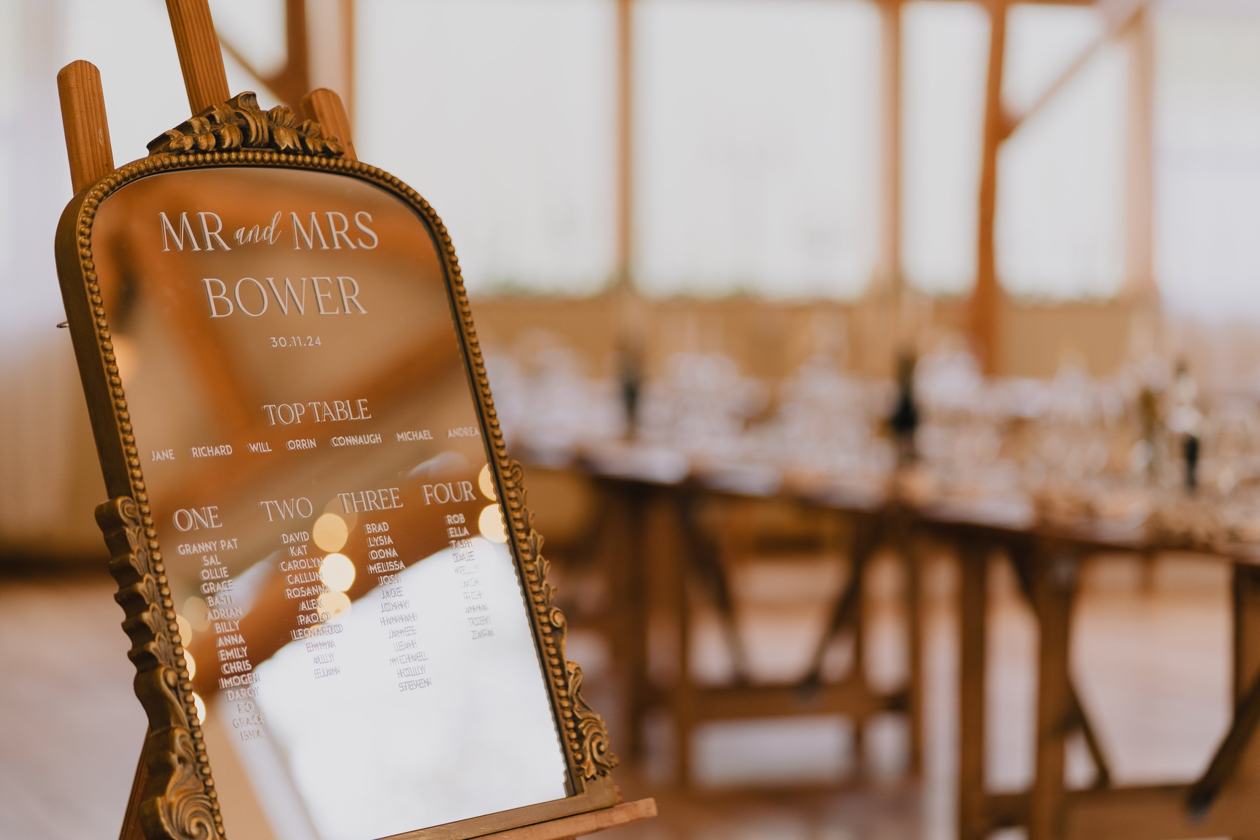 Wedding seating chart displayed on an ornate mirror with a wooden frame, showing the names of the bride and groom, date, and table assignments, in a warmly lit Owlpen Manor.